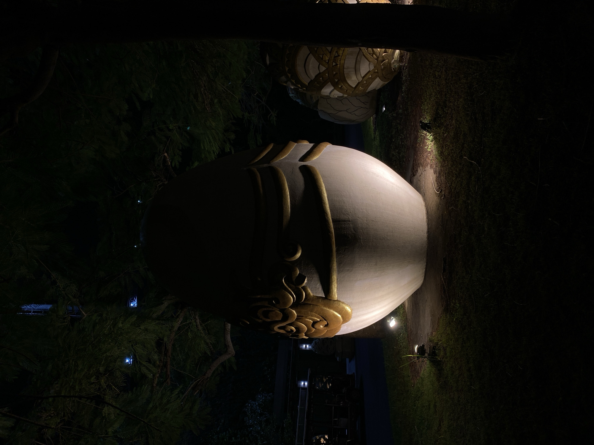 In Kuta Selatan, Indonesia, at night, a large, egg-shaped sculpture with gold ornamentation stands in a grassy area, illuminated by spotlights. The sculpture is light-colored with intricate, swirling gold patterns around its upper section. It is positioned in front of trees, with some of their branches extending into the frame. In the background, more sculptures are visible, also in an egg-like form and decorated with gold. A dark tree trunk is on the right side of the image. The overall atmosphere is dark, with the sculptures and some scattered blue lights in the trees being the primary sources of illumination.