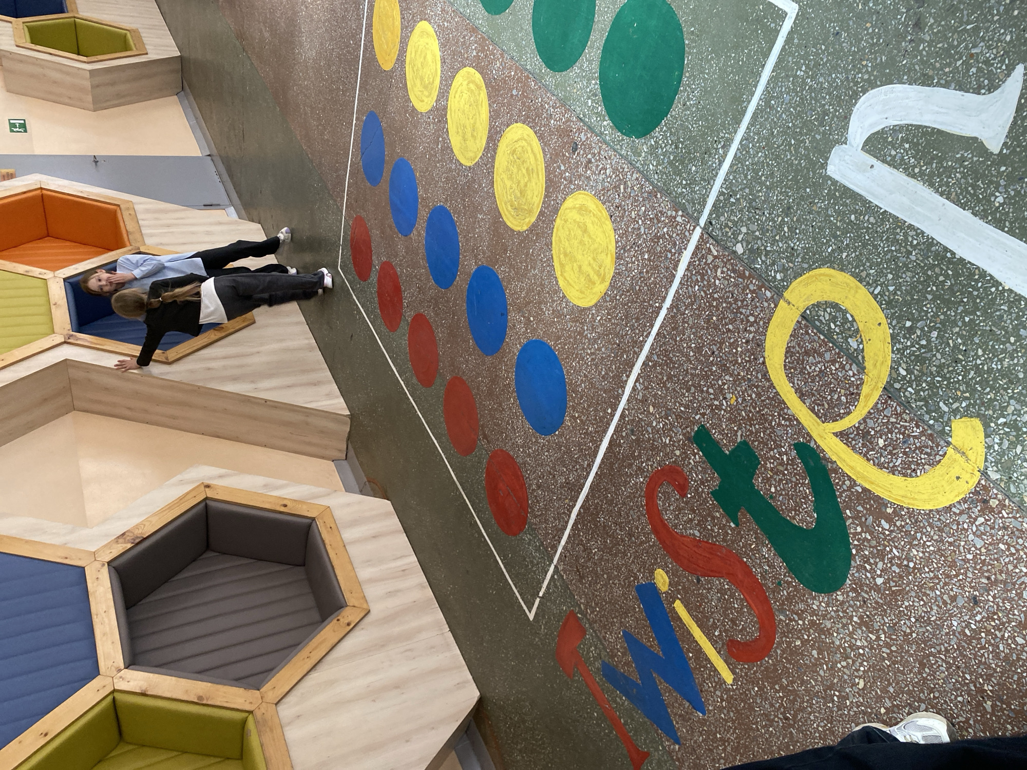 This image depicts an indoor recreational space in Votkinsk, Russia, featuring a giant, colorful Twister game painted directly onto the floor. The game area is a speckled terrazzo-like surface with rows of large yellow, blue, red, and green circles. The word "TWISTER" is also painted on the floor in large, stylized letters, each in a different vibrant color.
Two individuals, likely young people, are engaged in the scene. One person, wearing a dark top, dark pants, and white/purple shoes, is performing a plank position with hands on a red circle of the Twister game, actively playing. The second person, wearing a light blue long-sleeved shirt, dark pants, and white shoes, is positioned within one of the hexagonal seating cubbies, looking towards the camera with a slight smile, suggesting amusement or enjoyment.
The setting includes multiple hexagonal seating units with wooden frames and padded interiors in various colors like blue, orange, light green, and dark grey. These are integrated into the floor design, which transitions from the speckled game area to lighter, possibly wood-paneled surfaces around the seating. A small green and white sign, too indistinct to read, is visible on a wall in the background. The environment appears modern, designed for interactive play and relaxation.