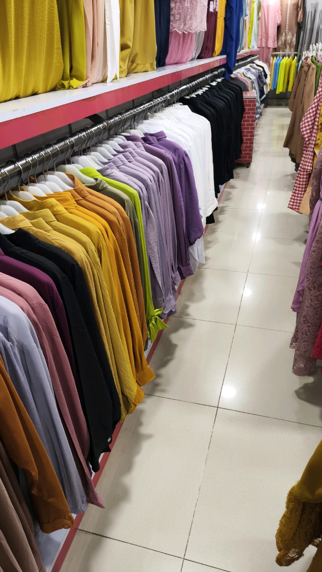 The image shows a clothing store in South Jakarta, Indonesia, specifically a retail display of women's clothing in a store. The foreground features rows of brightly colored shirts hanging on a metal rack, organized by color and ranging from shades of yellow and orange to purple and green. The shirts appear to be made of a lightweight fabric. In the background, more clothing items are visible, including dresses and other garments, displayed on shelves and racks. The store has a tiled floor and appears to be well-lit. The image is taken from a slightly elevated perspective, looking down at the clothing racks.