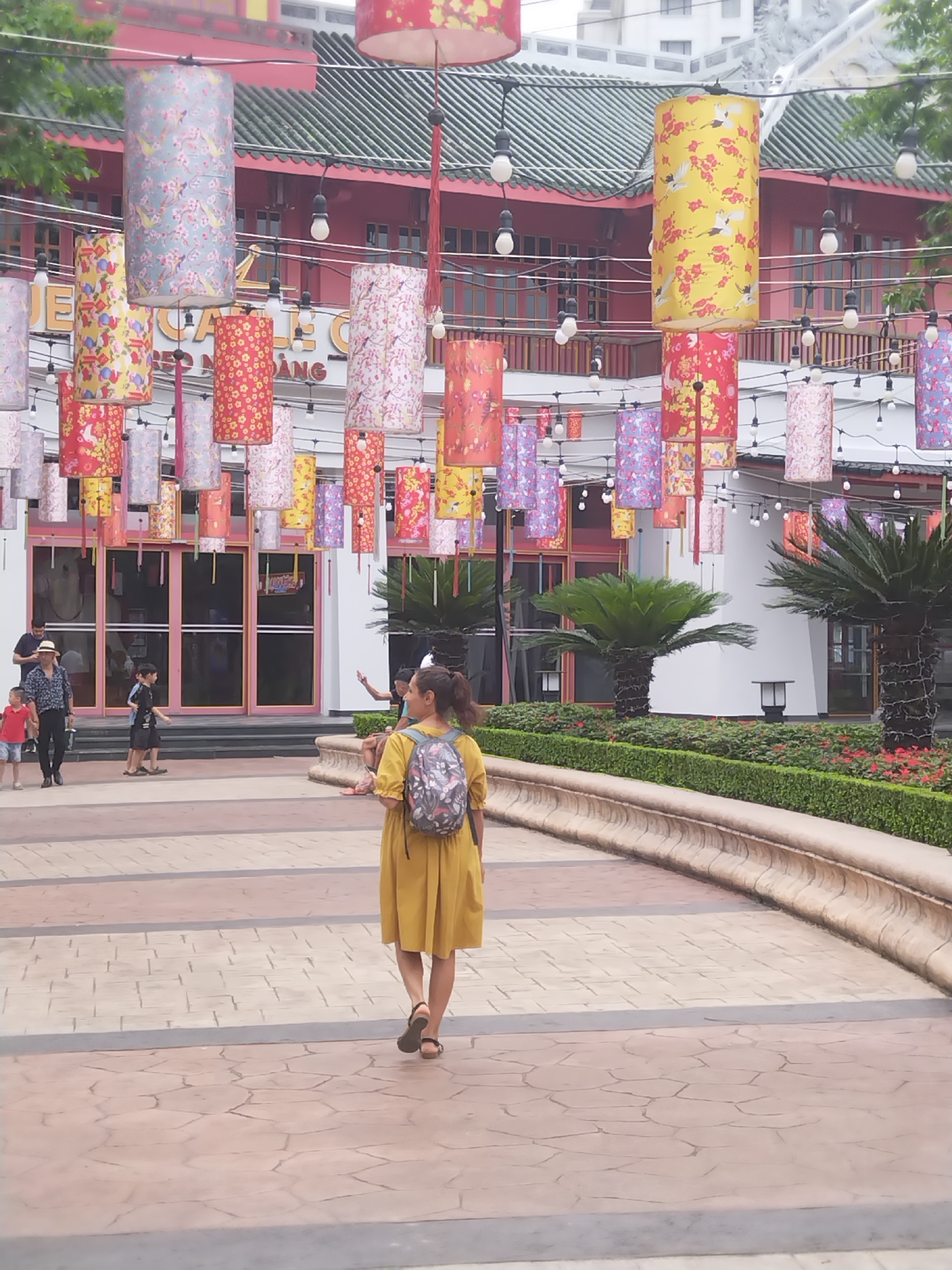 The image depicts a pedestrian area in Bai Chay Ward, Vietnam, decorated with numerous cylindrical lanterns hanging from overhead wires. The lanterns display a variety of floral and avian patterns. In the foreground, a woman wearing a mustard yellow dress and a backpack walks away from the viewer. In the background, several individuals walk towards a building with glass doors and windows. Landscaped greenery borders the walkway. Overhead string lights illuminate the area, accentuating the festive atmosphere. The background architecture features a red and green color scheme. The walkway is paved with light colored stone or concrete tiles.