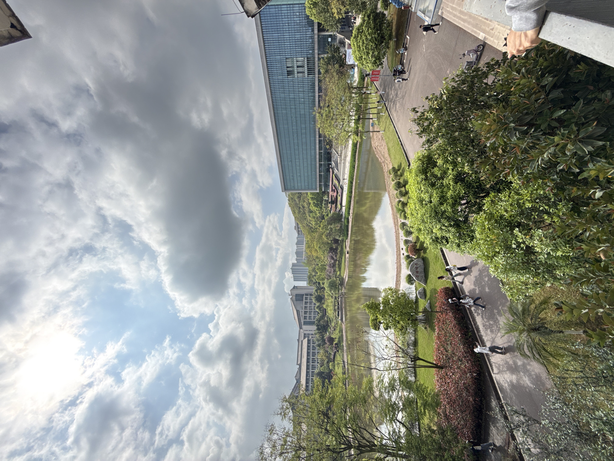 This is an eye-level outdoor shot of a cityscape under a cloudy sky, likely taken from an elevated position. The location is Jiangjin, China, suggested by the architecture and landscape.
In the middle ground, a canal or river reflects the sky and surrounding buildings. Along the canal, there are trees, shrubs, and grassy areas. The buildings include modern high-rises with glass facades.
On the right side of the frame, there is a paved walkway with several people walking along it. There are trees and shrubbery along the walkway. The top right corner of the shot includes a railing and part of a person's arm.