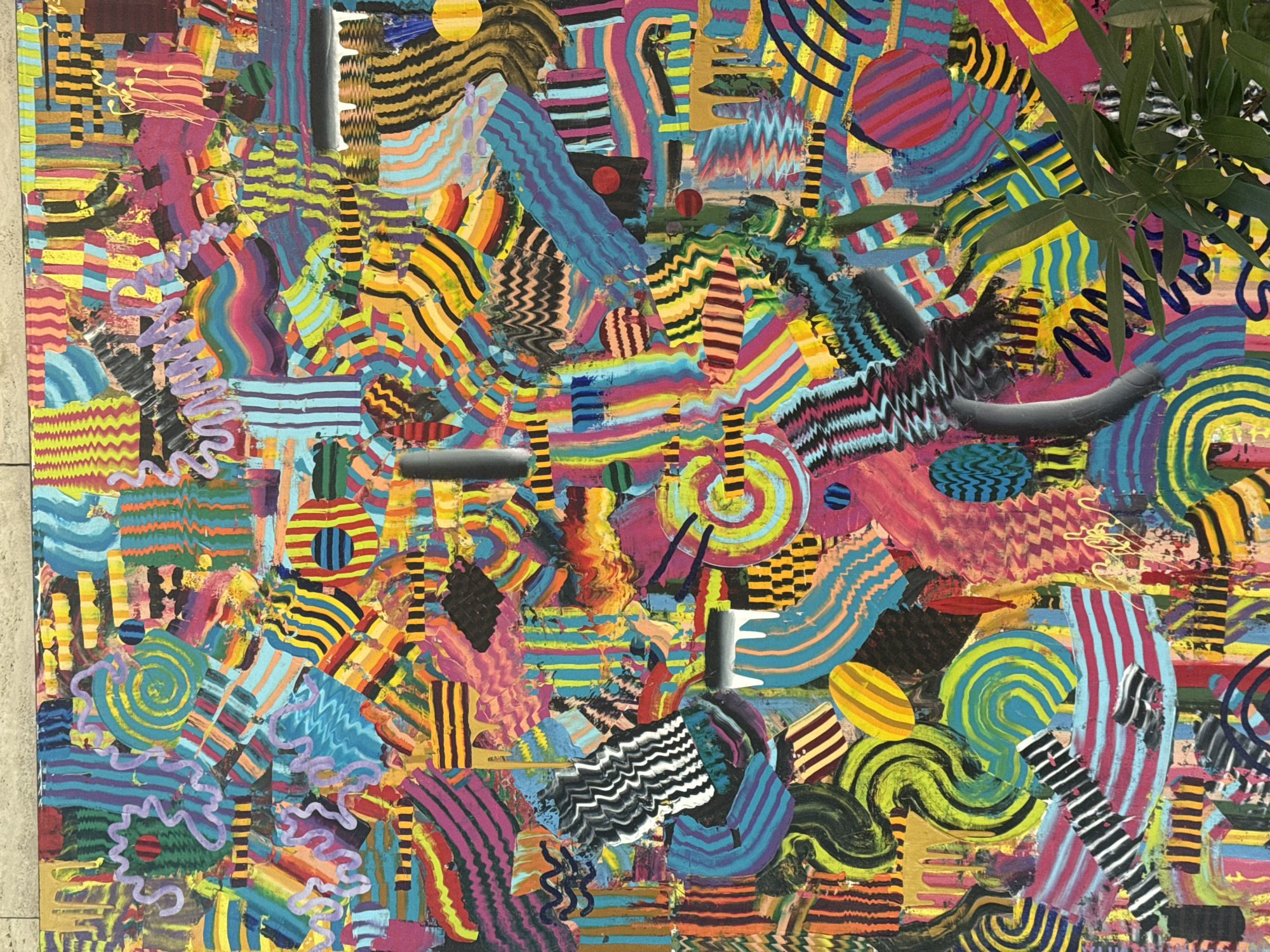 This is a detailed, full-frame, eye-level shot of a large, vibrant abstract painting. The artwork is densely packed with various geometric shapes, wavy lines, and textured patterns in a multitude of bright, contrasting colors, including blues, yellows, pinks, oranges, greens, and purples. The style is chaotic yet organized, with elements overlapping and interlocking. The painting appears to be made with a mix of mediums, possibly including acrylics and collage elements, given the varied textures.
The painting is displayed vertically. The top edge of the painting is against a textured, light-colored surface that appears to be a wall made of rectangular stone or concrete blocks. The bottom right corner of the painting is partially obscured by the foliage of a dark green plant, suggesting the artwork is displayed in an indoor environment, potentially a gallery, studio, or home, where plants are present.
There are no people, actions, or interactions visible in the image. The focus is solely on the artwork and its immediate surroundings.
Regarding the location in Belgrade, Serbia, there is no information available within the image itself to determine this. The scene described could be in any city with a gallery or artistic space.