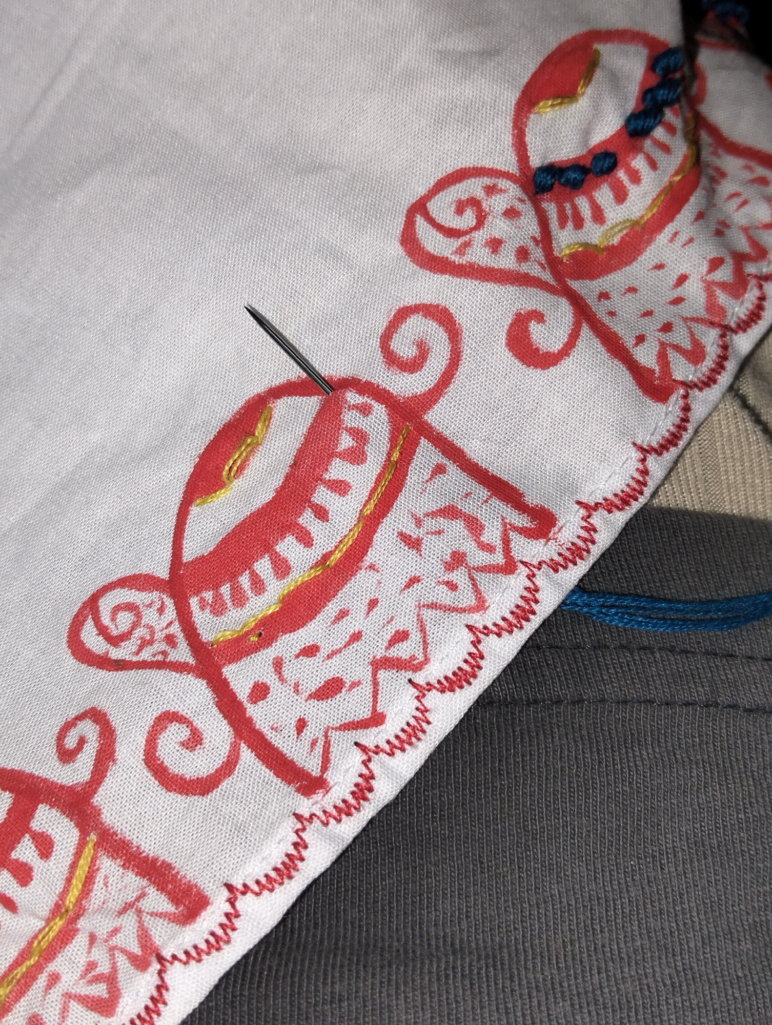 A close-up shows a white piece of fabric with red and gold embroidery lying on top of a gray piece of fabric. A needle is inserted into the white fabric. The style of the embroidery suggests South Asian origin. The image does not contain information to confirm the location as Dhaka, Bangladesh.