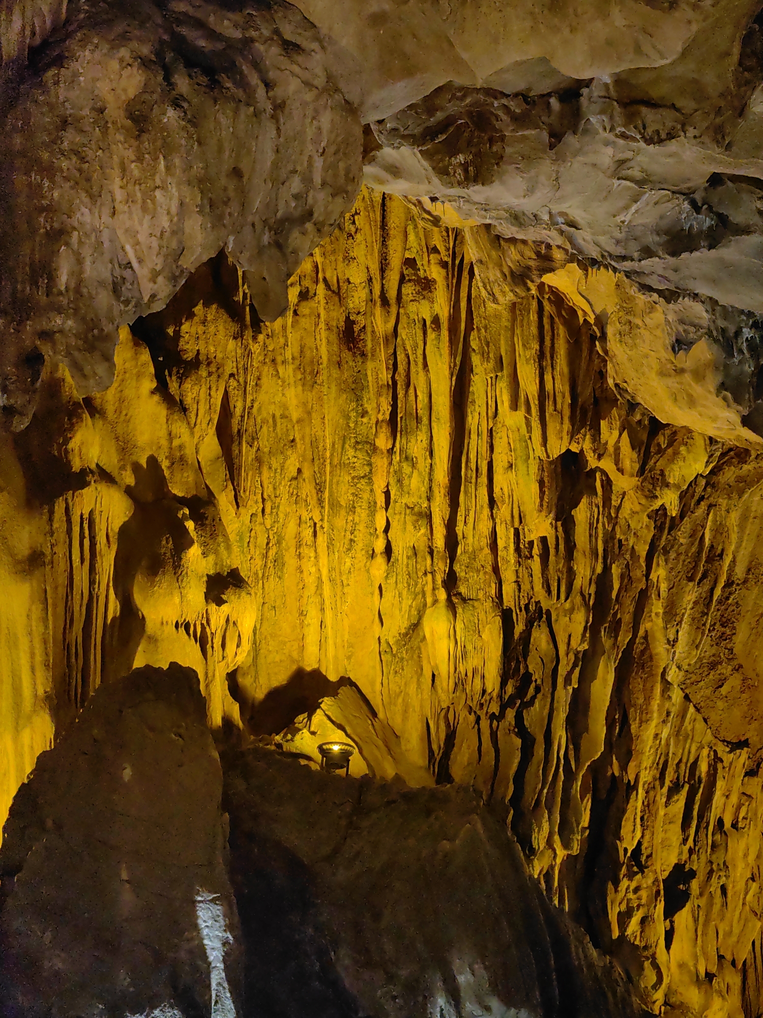 The image shows a section of a cave, presumably located in Lạng Sơn, Vietnam, as implied by the prompt. The cave's interior is illuminated with a warm, orange-yellow light. The primary feature of the image is a large rock formation with numerous stalactite-like structures hanging down. The rock appears rough and textured. At the base of the rock formations, there is a small lamp or light fixture perched on a relatively flat surface. The cave ceiling is visible in the upper portion of the image.