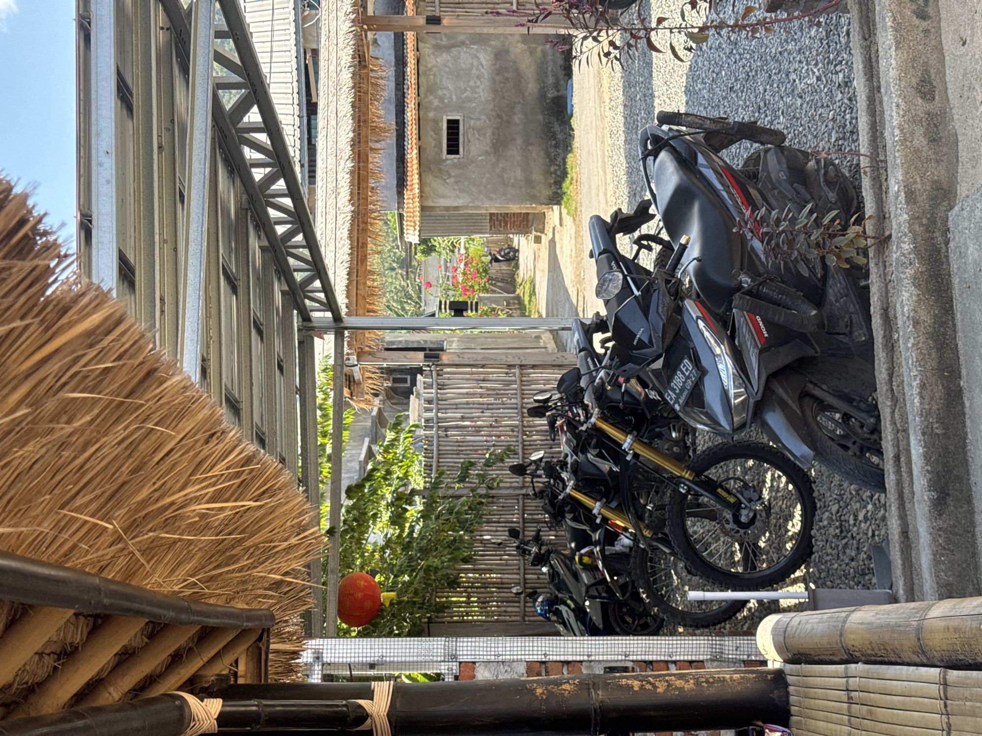 The image depicts a motorcycle parking area outside a building with a thatched roof and a metal awning in Kertasari, Indonesia. Several black motorcycles are parked on a gravel surface. A red lantern hangs from a bamboo pole. In the background, a building with a grey concrete exterior and an orange tiled roof is visible, along with a wooden fence and greenery. The scene appears to be outdoors in daylight, with a clear blue sky overhead. No people are present in the image.