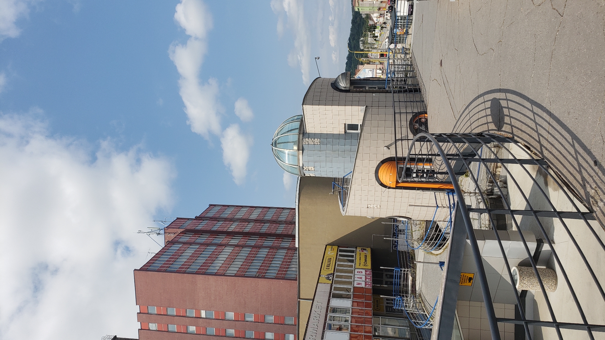 The image captures a view of the "Prima" shopping center in Prešov, Slovakia, on a sunny day. The architecture is modern, featuring a tall, red brick building with many windows on the left, and a more rounded, multi-colored structure with a glass dome on top in the center. A curved metal railing forms a walkway or balcony in the foreground, casting a distinct shadow on the cracked concrete path. The background shows a street with cars and buildings, suggesting an urban setting. The sky is blue with scattered white clouds. There are no visible people in the image.