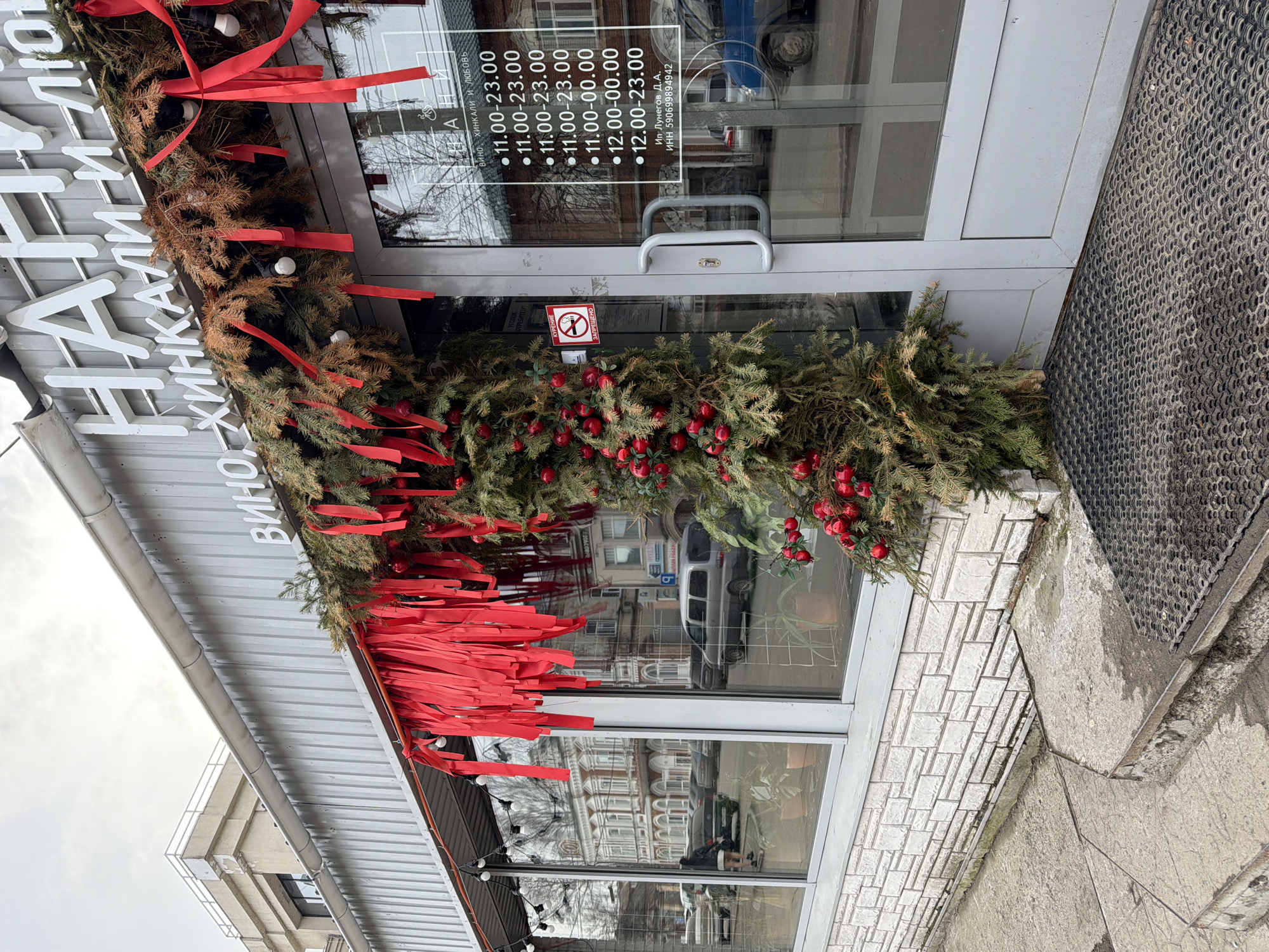 Here's a description of the image:
The image shows the entrance to a business, possibly a restaurant or bar, in Votkinsk, Russia. The sign above the entrance says "Хинкали и любовь" which translates to "Khinkali and Love" (Khinkali is a type of Georgian dumpling). It also mentions "ВИНО," which translates to "wine." The entrance is decorated with a garland of evergreen branches, red ribbons, and red berries. The door is glass, and visible through the glass are the reflection of surrounding buildings, and an opening hours sign indicating the business is open from 11 AM or 12 PM to 11 PM or 12 AM. There is also a "No Smoking" sign on the door. A gray mat lies on the ground in front of the entrance. The building's facade is white brick. A partial view of neighboring buildings and the sky is also visible.
