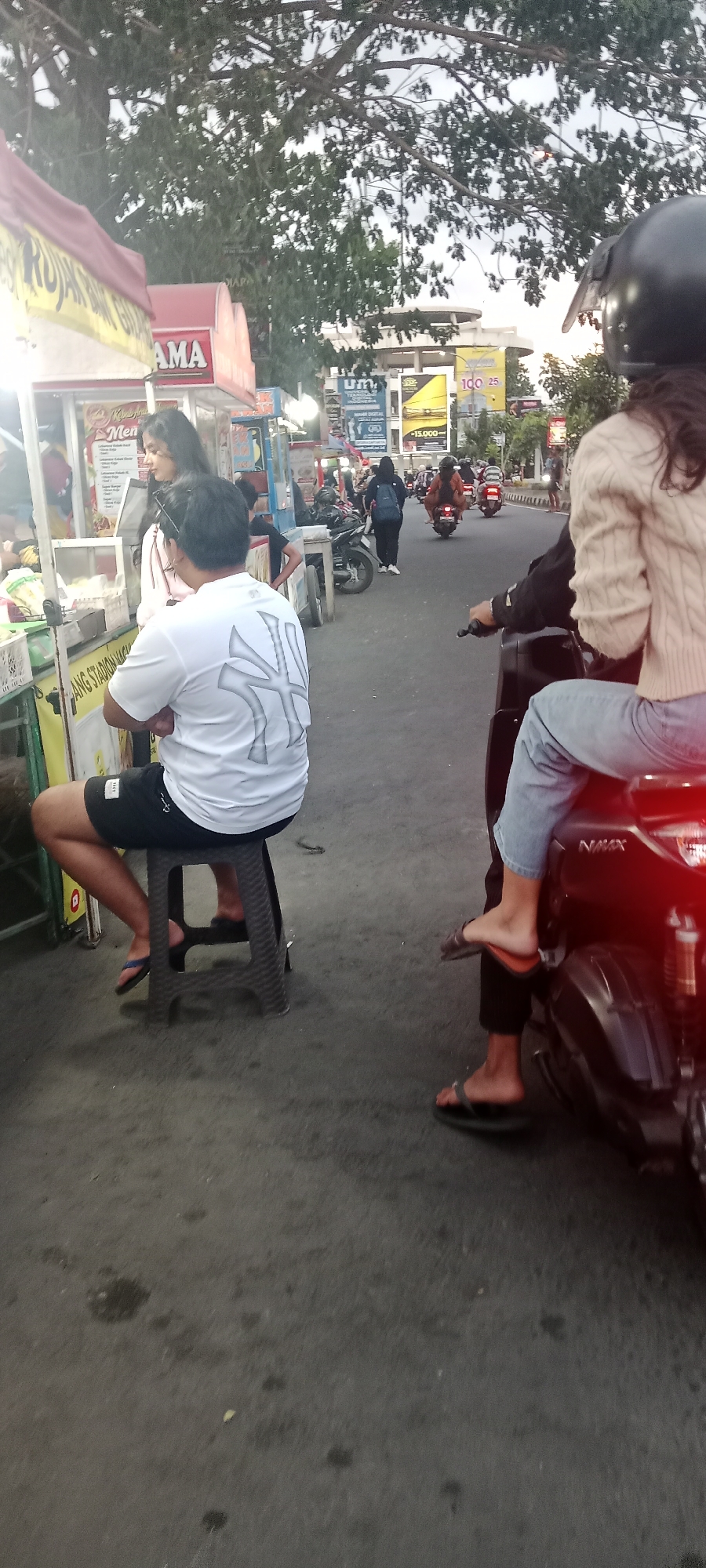 In the city of Ngemplak, Indonesia, on a street with a grey asphalt surface, a man wearing a white t-shirt with a large grey graphic and black shorts sits on a dark grey plastic stool. He is facing away from the camera and towards a food stall. To his left, a woman with long dark hair, wearing a white top, stands near the food stall, looking in the same direction. In the background, several people are on motorcycles. One person, wearing a black helmet and a light brown knitted sweater over a dark shirt, is riding a dark motorcycle. Another person, wearing a light brown top and blue jeans, is a passenger on the back of the motorcycle, also facing away from the camera. Further in the background, a group of motorcycles are driving down the street. The environment appears to be an outdoor market or street food area, with the food stall prominently featured on the left side of the image. Trees with dense green foliage are visible overhead, and various signs and advertisements are present in the background. The lighting suggests it is either early morning or late afternoon.
