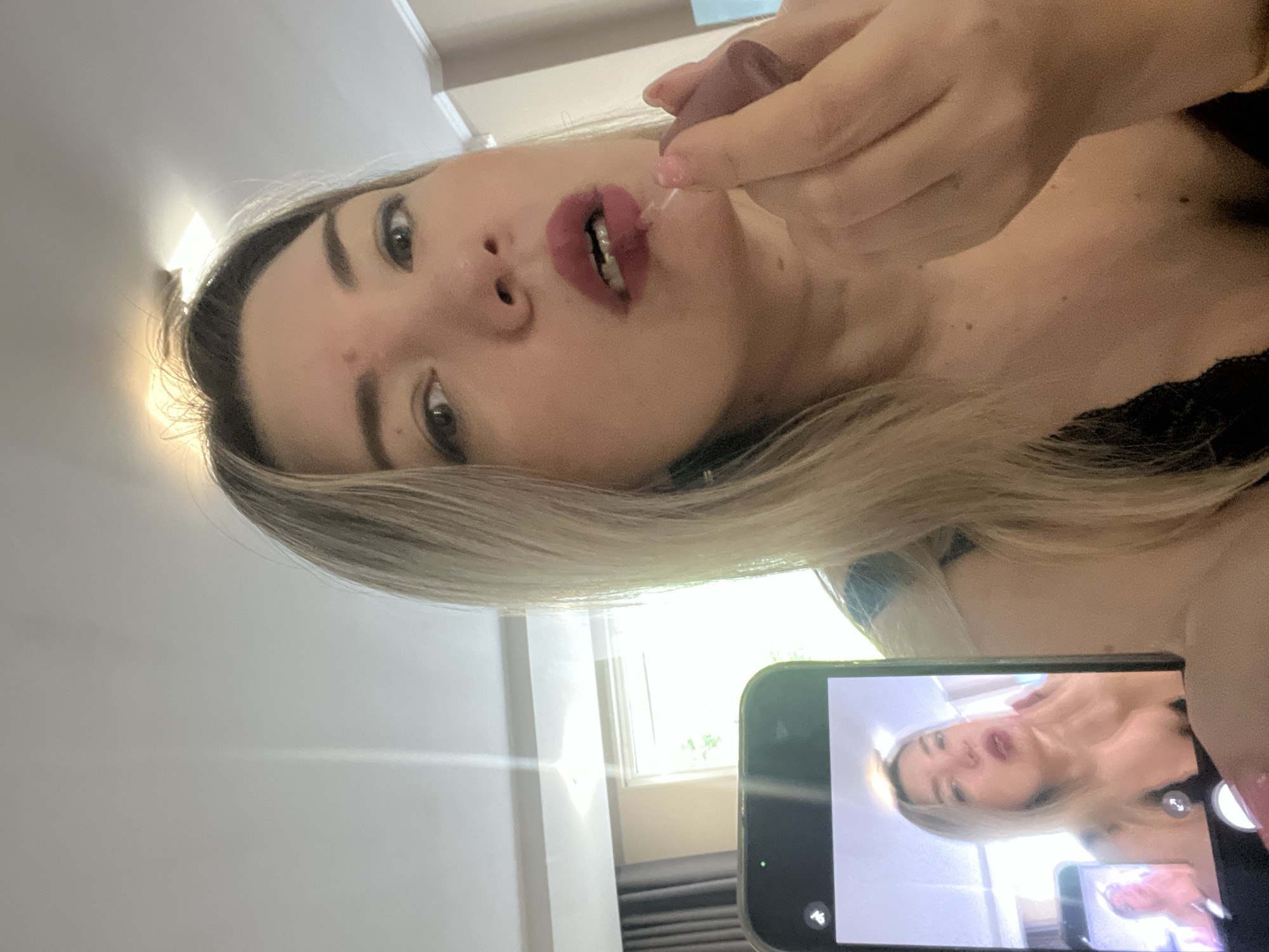 A woman with blonde hair applies dark pink lipstick to her lips. She is holding a makeup applicator in her right hand. In the foreground, a smartphone displays a selfie of the woman applying lipstick, with a smaller inset image of herself applying makeup. The background appears to be an indoor setting, possibly a room in a hotel, with curtains and a window visible. There is no indication of the city of Sochi, Russia, in the image.