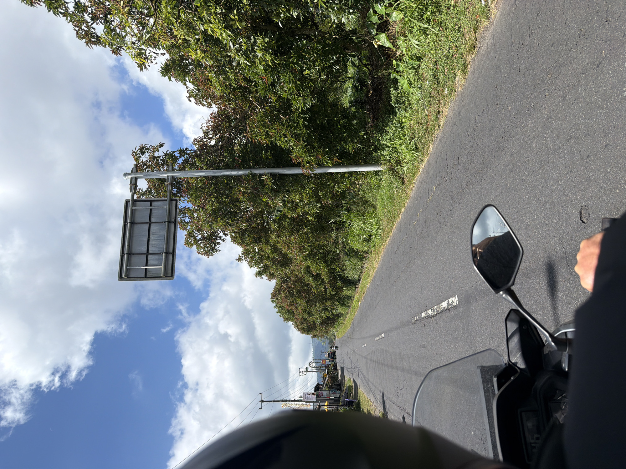 The image depicts a point-of-view shot from a motorcycle traveling on an asphalt road in Kintamani, Indonesia. The road is lined with lush green trees and vegetation on the right side, and there are buildings and power lines on the left. A large, empty billboard structure is mounted on a pole on the right side of the road. The sky is blue with scattered white clouds. In the distance, a few vehicles and a motorcyclist are visible. The rider's hand is on the handlebar, and their helmet is in the foreground, obscuring part of the view. The scene appears to be a rural or semi-urban setting, typical of Indonesian roadways.