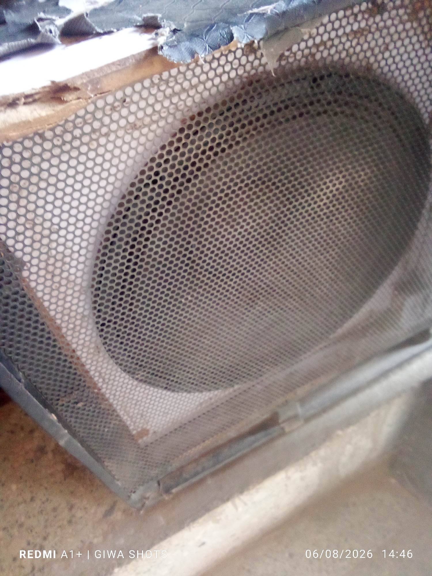 The image shows a close-up of a damaged speaker. The speaker is housed in a wooden cabinet, part of which is covered with a metal grille. The grille has a pattern of small, circular holes. The top of the cabinet is torn and frayed, revealing the material underneath. The speaker cone itself is visible through the grille, appearing dusty and somewhat faded. The overall condition of the speaker suggests it is old and has been exposed to the elements or rough handling. The text overlay indicates the device used to take the photo is a REDMI A1+ and the location is GIWA SHOTS, with the date 06/08/2026 and time 14:46. The scene appears to be indoors, or at least in a sheltered area, given the lighting and the lack of harsh outdoor elements, though the dirt and damage suggest it might have been stored or used in a less-than-ideal environment. There are no people visible in the image.