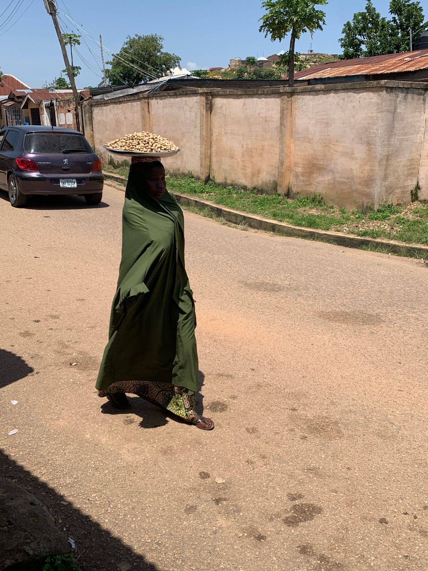 A woman walks down a paved street in Jos, Nigeria, carrying a large tray of peanuts on her head. She is wearing a dark green abaya and patterned trousers. A dark purple Peugeot car is parked on the left side of the street. A concrete wall runs along the right side of the street, with some vegetation growing at its base. The sky is clear and blue, with a few trees visible in the background. The sun is casting shadows on the street.