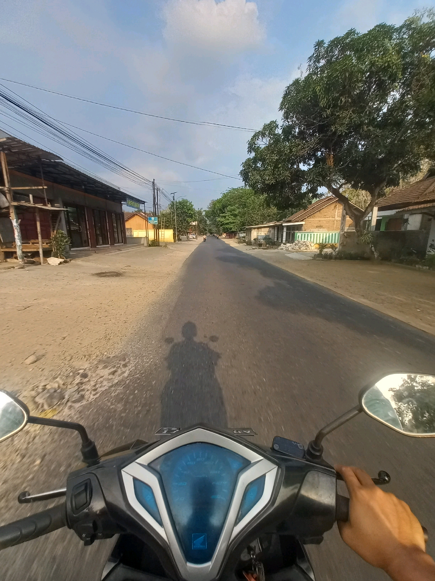 This is a first-person perspective, forward-facing, medium shot video of riding a motorcycle on a paved road in Ngarjosari, Indonesia, at what appears to be dusk. The road is flanked by a dirt shoulder on the left and a sandy shoulder on the right, leading to buildings and trees. On the left, there's a building with a corrugated metal roof, under construction with wooden scaffolding. Further down the road on the left are more shops. On the right, there are houses with tiled roofs and a large tree. The sky is partly cloudy with a clear blue sky. The motorcycle's handlebars and dashboard are visible in the foreground. A human hand is on the right handlebar. The shadow of the motorcycle and rider is cast on the road. The environment suggests a rural or suburban setting.