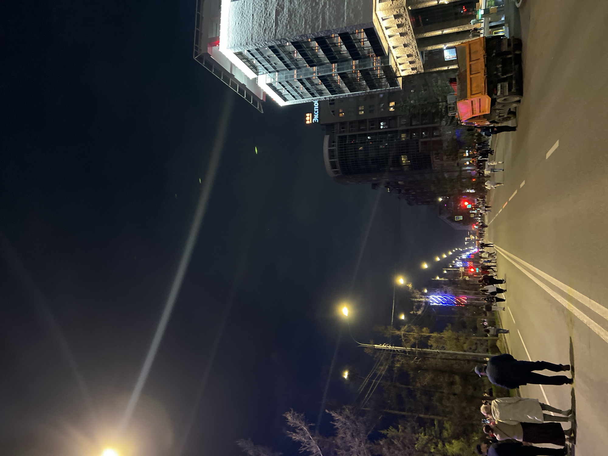 The image depicts a street scene in Krasnoyarsk, Russia at night. The "ЭКСПО" (Expo) building is visible. A crowd of people is present on the street, some appearing to be standing and observing, while others are walking. A large yellow truck is parked on the side of the road. Streetlights illuminate the scene and buildings. Trees line the street.