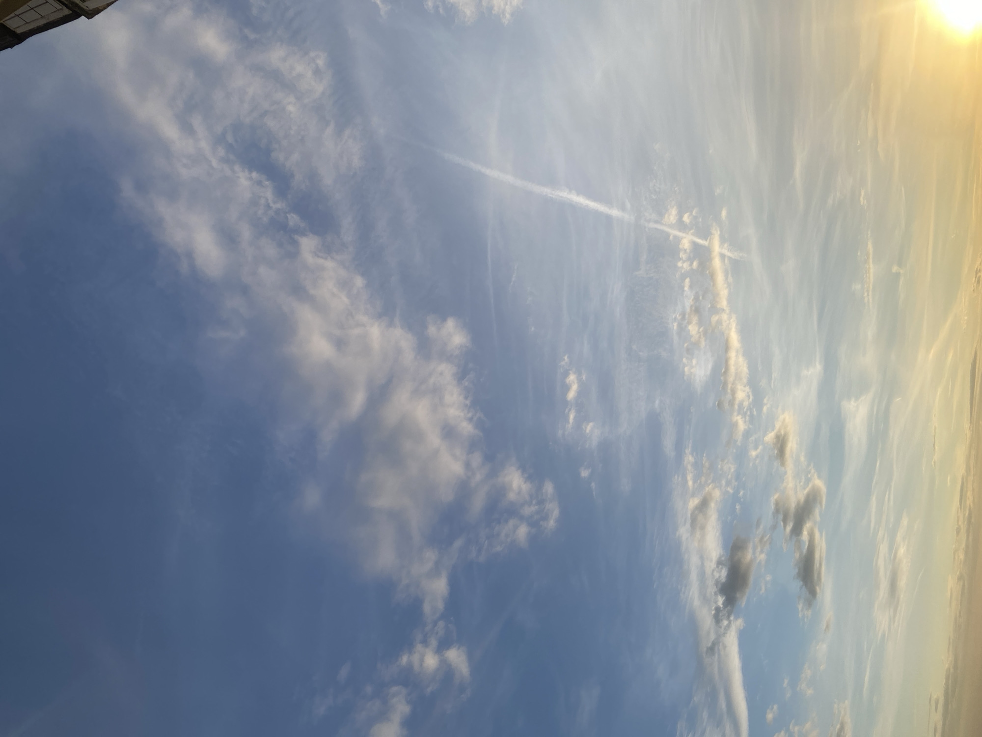 The image shows a blue sky with scattered clouds. There is a contrail from a plane visible in the upper center of the image. The sun is visible in the upper right corner, creating a bright light. The city location is unknown.