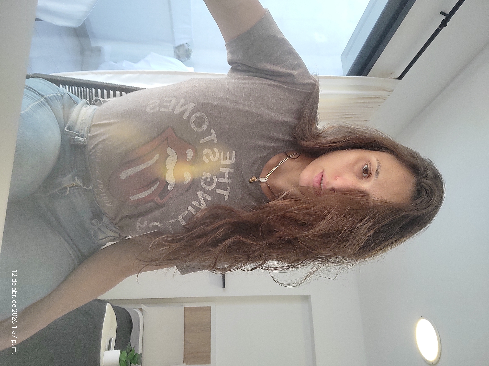 A woman wearing a gray Rolling Stones t-shirt and blue jeans is posing indoors. She is wearing a gold necklace. The room is painted white and has a window with white curtains. A partially visible piece of furniture and a light fixture can be seen. The photograph includes the date "12 de abr. de 2026 1:57 p. m.". The location, Buenos Aires, Argentina, is not evident in the image.