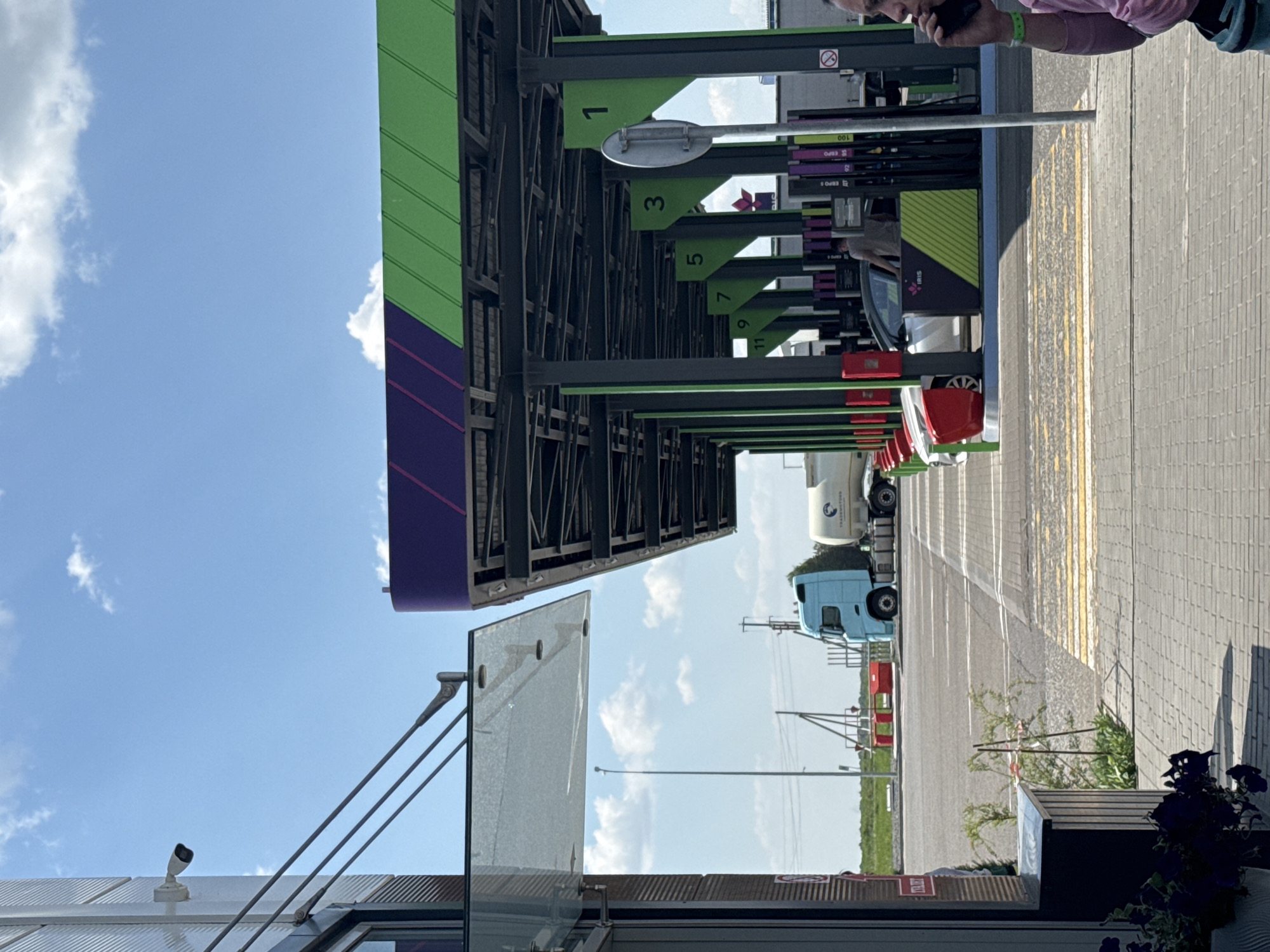 The image depicts a gas station in Bol'shie Nyrsi, Russia. Gas pumps numbered 1 through 11 are visible under a green and purple canopy. Several vehicles, including cars and a light blue semi-truck with a white tanker, are parked at the pumps. The ground is paved with grey brick and marked with yellow lines. Above and to the left of the gas station canopy is a light blue sky with scattered clouds. A security camera is attached to a building in the foreground. The bottom right of the image shows a planter with dark purple flowers. An individual is partially visible on the right edge, using a mobile phone.
