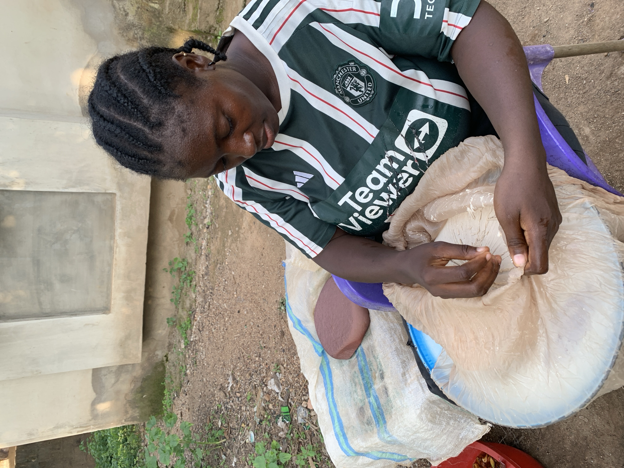 An overhead medium shot captures a dark-skinned woman with braided hair sitting in a purple plastic chair outdoors in Akwanga, Nigeria. She is wearing a green and white striped Manchester United soccer jersey with the TeamViewer logo and the Manchester United crest visible. The woman is looking down at her hands, which are sifting through a tan plastic bag filled with a white powder. To her left, on a woven mat, are a brown, rounded object and a blue bowl. The background consists of a rough, tan concrete building with a small window and overgrown vegetation. The ground is covered in dirt and small stones.