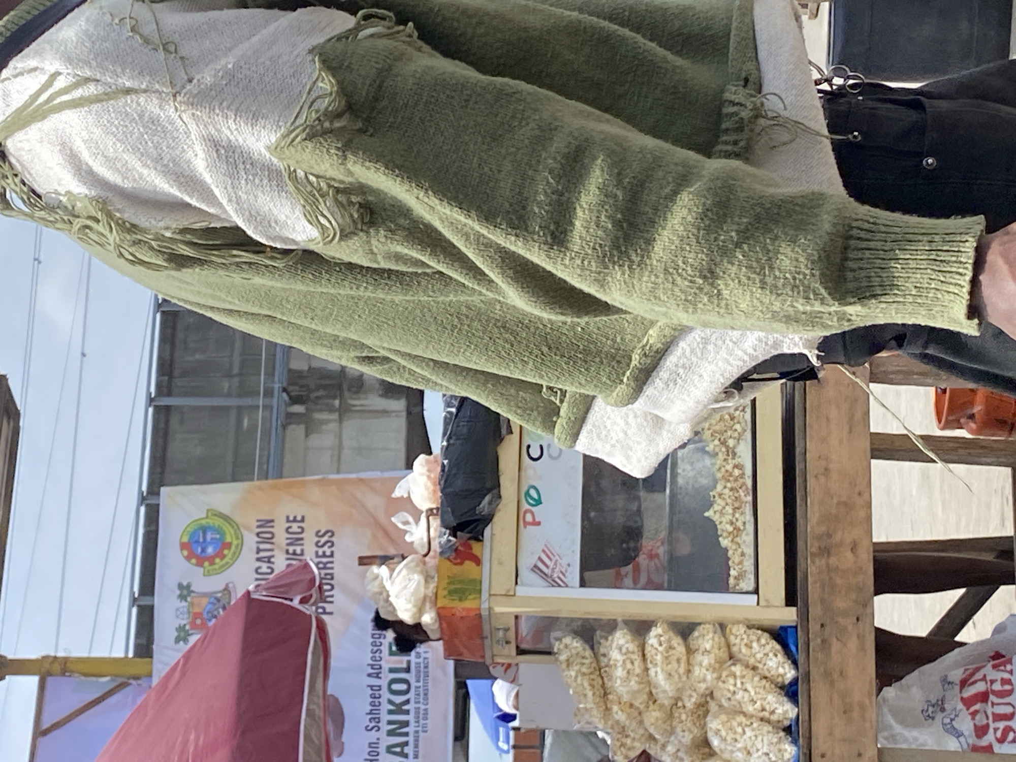 The image shows a person wearing a green sweater over a white top. The person is positioned near a popcorn stand in what appears to be Lagos, Nigeria. The stand includes a sign that reads "POC", and it has bags of popcorn on display. Adjacent to the stand is a banner promoting Hon. Saheed Adeseg ANKOLI, a member of the Lagos State House of Assembly for Eti Osa Constituency 1, and discussing "Education Science Progress."