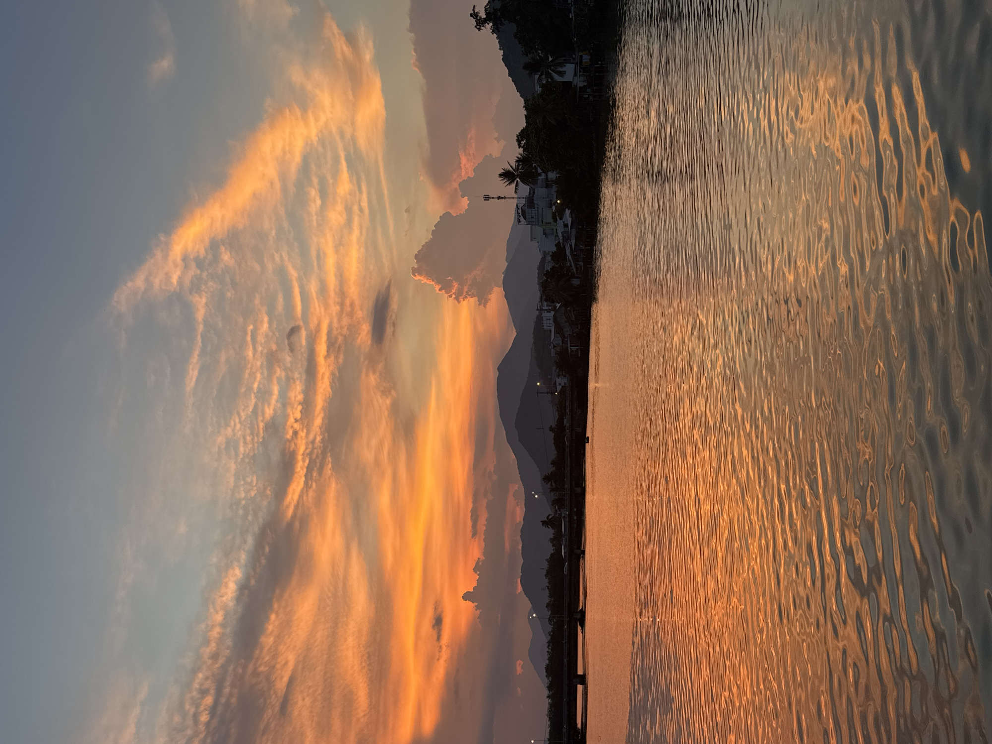 The image depicts a serene river scene in Nha Trang, Vietnam, during a breathtaking sunset. The sky is a canvas of vibrant orange and pink hues, with wispy clouds creating dramatic patterns. A bridge spans the river in the distance, with a few lights visible on its structure. Along the riverbank, houses and lush green trees dot the landscape, with mountains forming a majestic backdrop. The water's surface reflects the fiery colors of the sky, creating a shimmering, golden expanse. The overall atmosphere is one of tranquility and natural beauty.