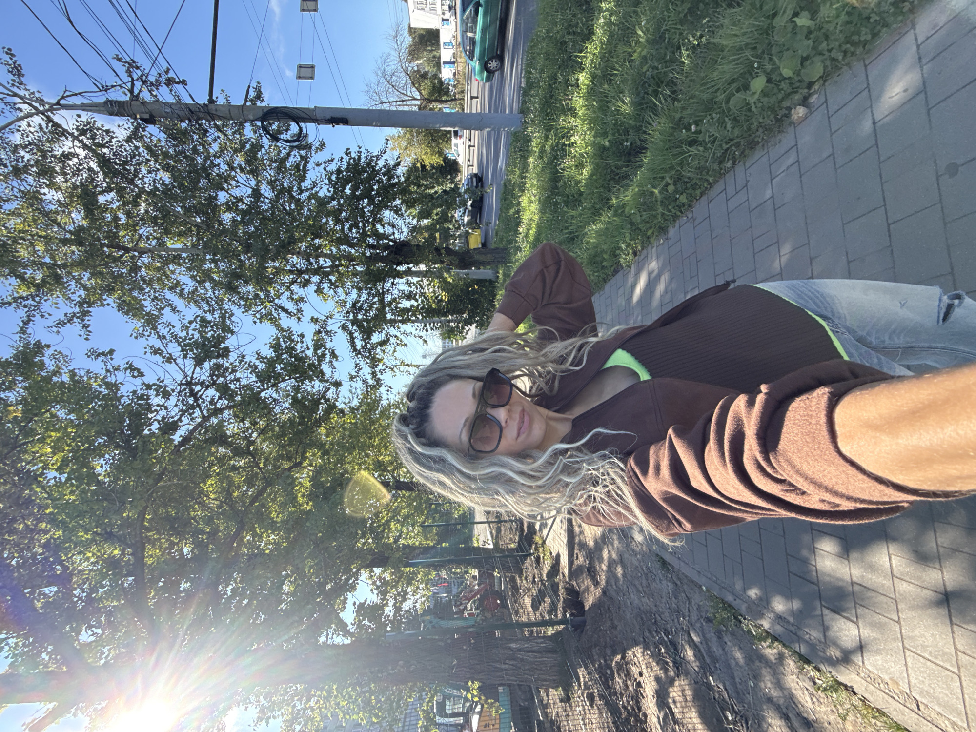The image shows a woman with long, curly blonde hair and sunglasses taking a selfie on a sidewalk. She is wearing a brown zip-up jacket over a neon green top and distressed blue jeans. To her left, there is a fenced-off construction area with a yellow excavator. To her right, there is a grassy area and a road with cars passing by, including a green car. Trees line the sidewalk and road, and utility poles with wires are visible in the background. The sun is shining brightly, casting shadows and creating lens flare. The location is identified as Zhukovo, Russia.