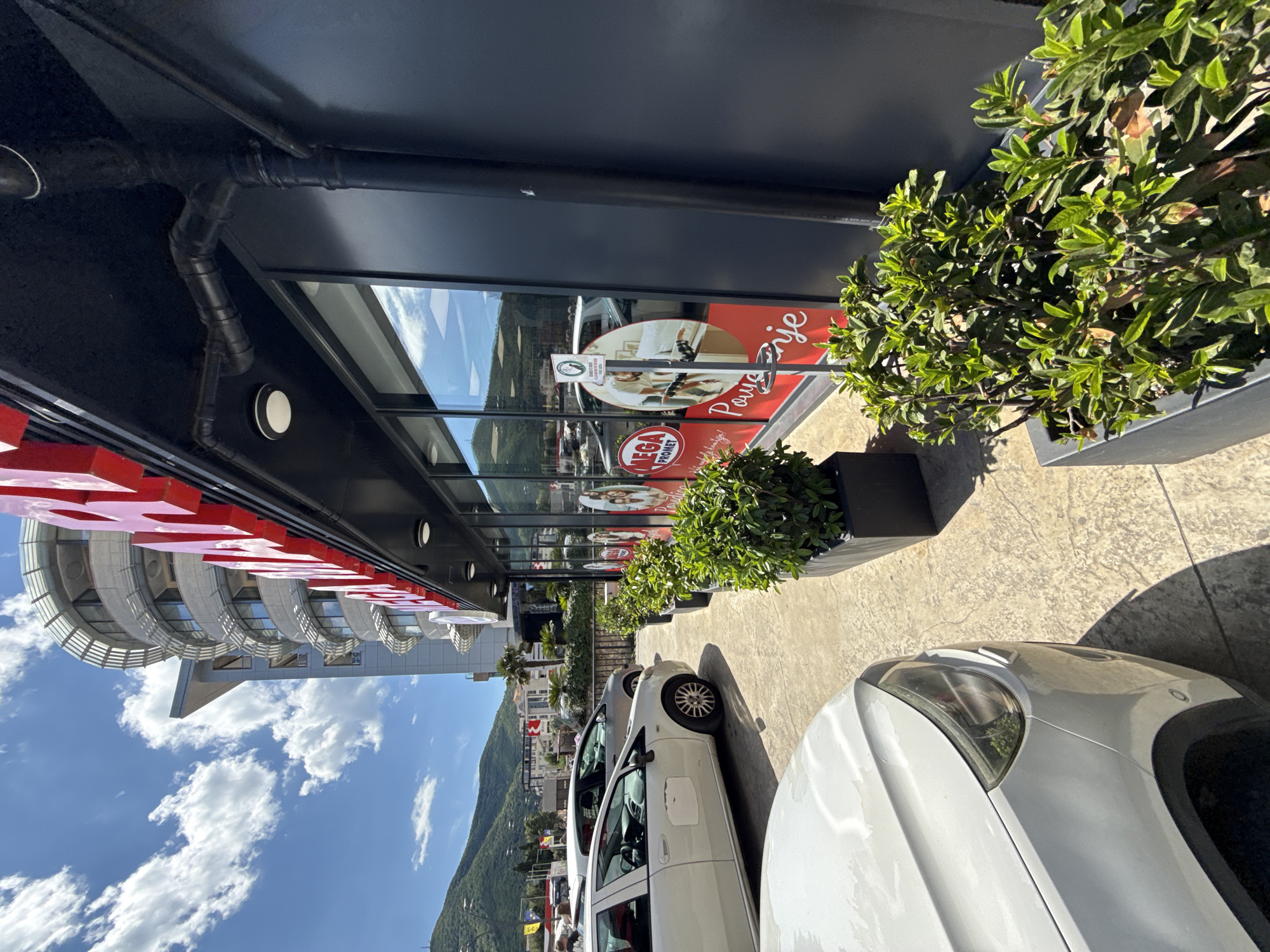 This is a vertical, medium-angle shot of a row of parked cars in front of a modern building with large windows. The building's facade is dark, with a row of red lettering across the top, spelling out "MEGA PROMET". A black pipe system runs along the underside of the building's overhang. Green potted plants are strategically placed along the sidewalk in front of the building.
In the foreground, the front of a white car is visible, with its headlight and part of the hood in focus. Behind it, another white car and a gray car are parked. The sidewalk is paved with a textured concrete material, and shadows from the building and plants indicate it is daytime. The background shows a slightly overcast sky with scattered white clouds, and a lush green mountainside. The scene appears to be an outdoor commercial area, possibly a shopping center or a row of businesses.
Reflected in the building's windows, a circular advertisement features people and text. One prominent sign on the building reads "Pivrije". The overall impression is a sunny day in a commercial district.