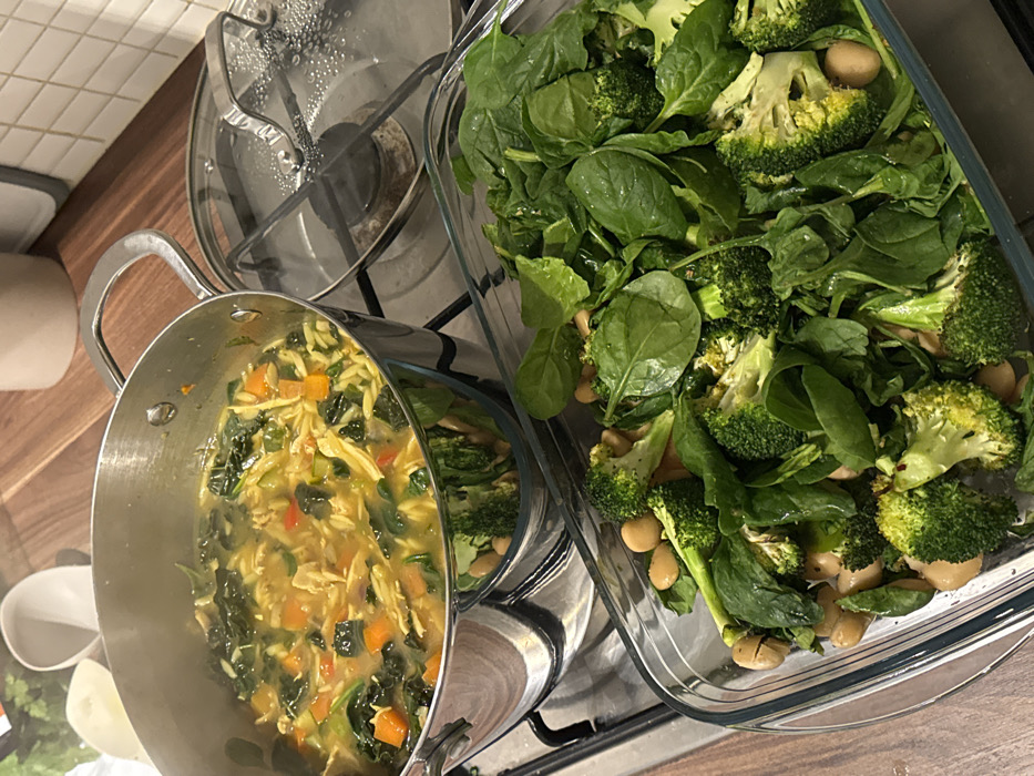A metal pot filled with soup containing pasta, chopped carrots, and leafy greens sits on a stovetop. Beside it, a clear glass dish holds a salad mix of spinach, broccoli florets, and white beans. A glass lid rests on a nearby burner. The scene suggests a healthy meal being prepared in a kitchen setting.