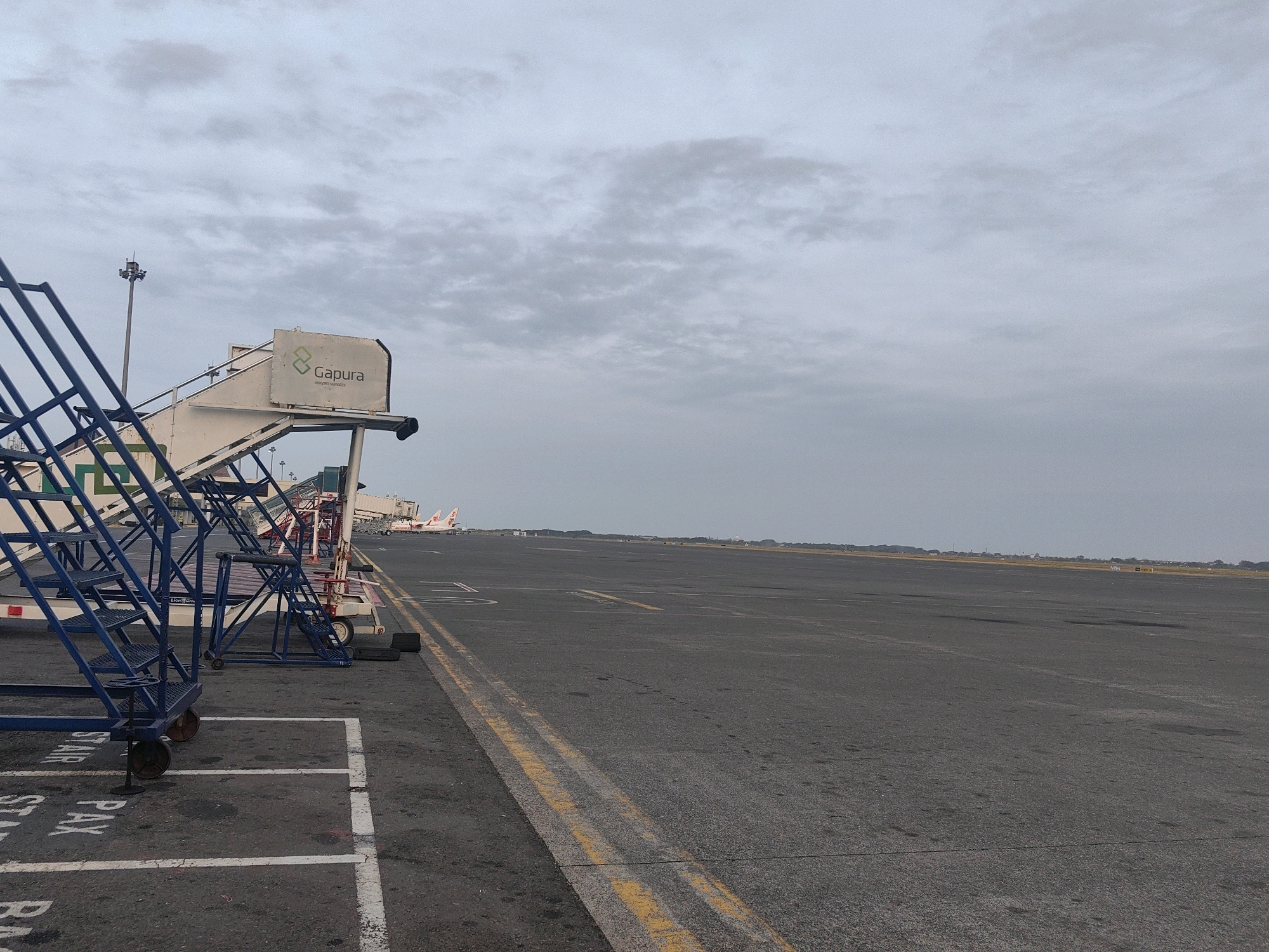 The image shows an outdoor scene at an airport, possibly at dawn or dusk due to the overcast sky. In the foreground, a blue metal mobile airstair is positioned on the tarmac, with white lines painted on the asphalt indicating parking or queuing areas. To the left, another similar airstair is partially visible. Behind the airstairs, a larger, white mobile boarding bridge with the logo "Gapura" is connected to a building, presumably an airport terminal. In the middle ground, several commercial airplanes are parked on the tarmac, some with their logos visible, suggesting an active airport environment. The background consists of a long, empty asphalt runway stretching towards the horizon, with sparse vegetation and buildings visible in the far distance. The sky is filled with heavy, grey clouds. The location is identified as Segoro Tambak, Indonesia, a coastal area known for its fishing villages and its proximity to the Juanda International Airport in Surabaya.