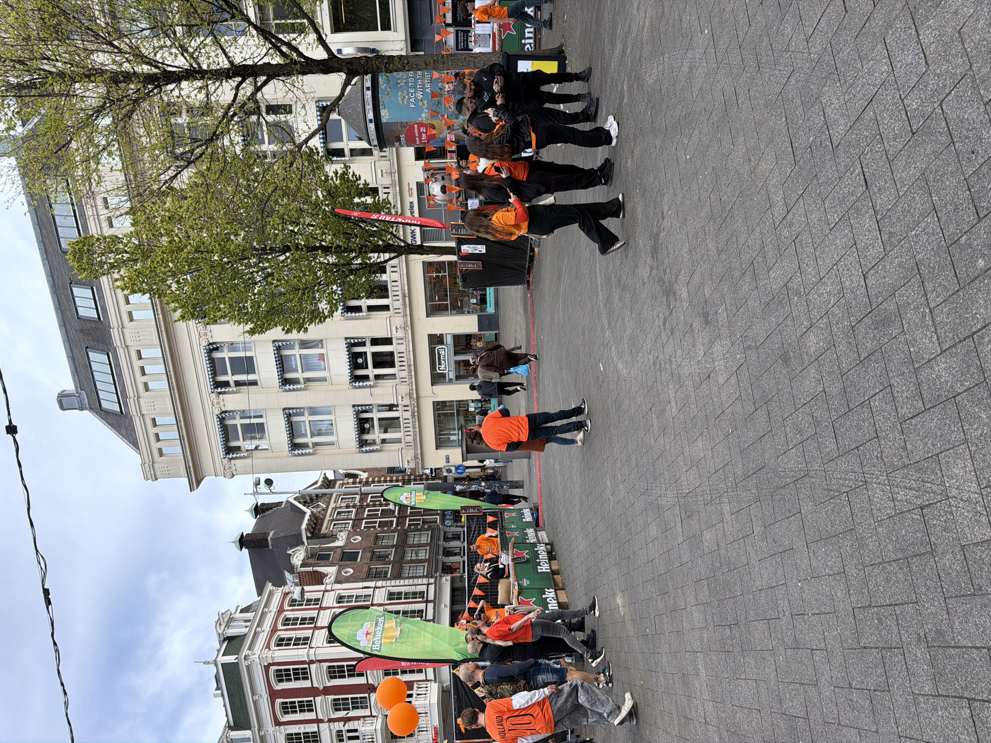 This image depicts an outdoor urban scene in Amsterdam, Netherlands, characterized by a gathering of people, many dressed in orange, suggesting a festive event. The setting is a paved square surrounded by multi-story buildings showcasing traditional Dutch architecture, some with distinct gabled roofs and large windows.
Numerous individuals are visible throughout the square. Many are wearing bright orange tops, some featuring "HOLLAND" and the number "10" on their backs, a strong indicator of a national celebration like King's Day. Green and orange inflatable banners bearing the "Heineken" logo are prominent features, along with stacks of "Heineken" boxes and orange flags strung across the area. Two orange balloons are also visible in the foreground.
Activity appears to be casual socialising; people are standing, walking, and gathering in groups. A large poster on a building in the background features text like "FACE TO FA WITH THI ARTIST" and "Sook online 3 for 2!", suggesting an art or retail promotion. Other visible text includes "GWKS velex", "Normal", and "eink".
The time of day is daytime, under an overcast sky. Trees scattered around the square are in full leaf, indicating spring or early summer. The ground is paved with rectangular grey stones arranged in a herringbone pattern. The prevalence of orange, the "Heineken" branding, and the "HOLLAND" text strongly point to a celebration tied to Dutch national identity.