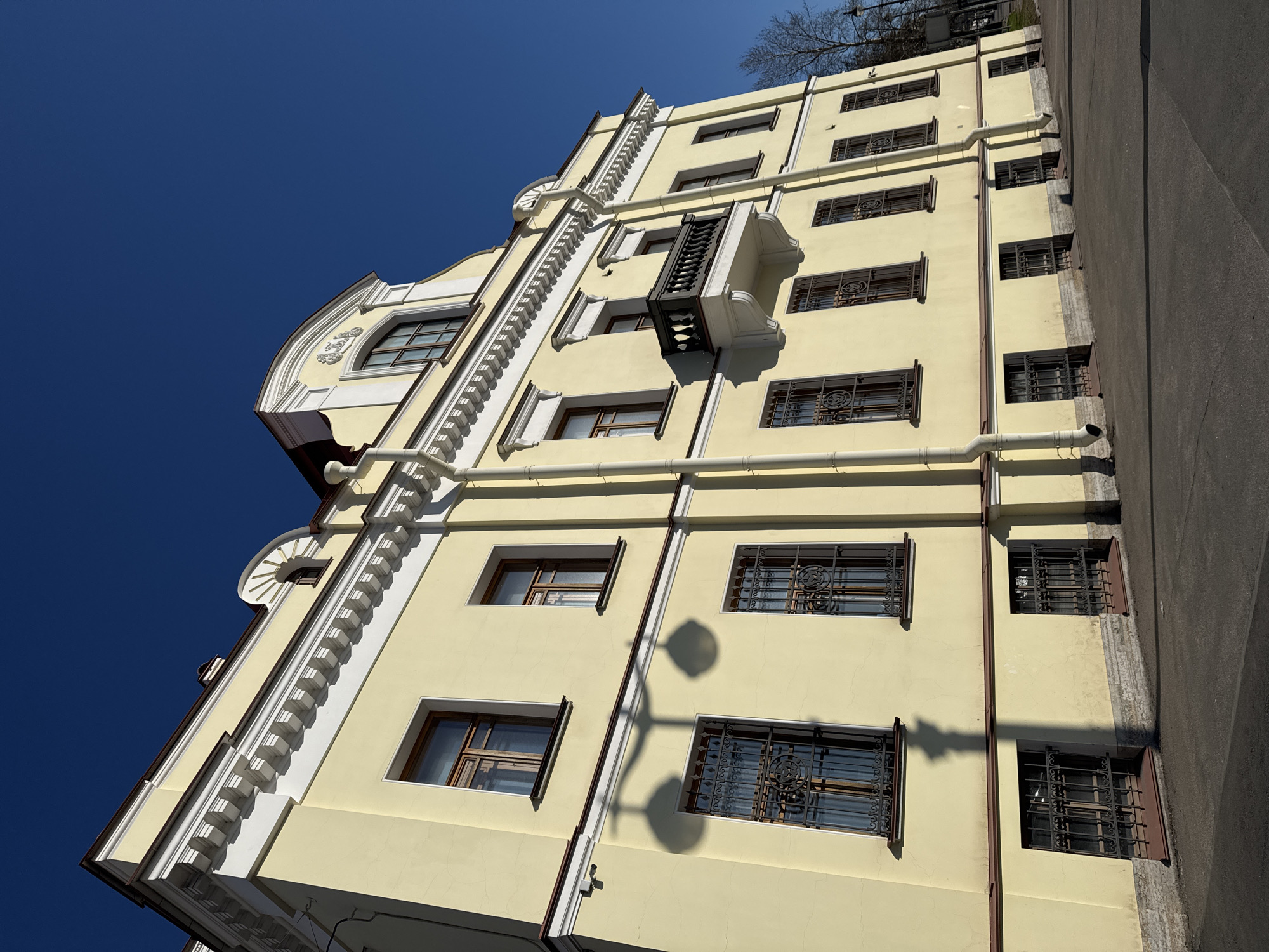 The image depicts a low-angle view of a multi-story building in Saint Petersburg, Russia. The building's facade is light yellow with white architectural details. Windows are visible, some with decorative iron grilles. A small balcony protrudes from the building. Rain gutters run vertically down the building's side. The sky is a clear, deep blue. A portion of an asphalt road is visible on the right side of the frame. Shadows from a street lamp are cast on the wall. Bare tree branches can be seen against the sky in the background.