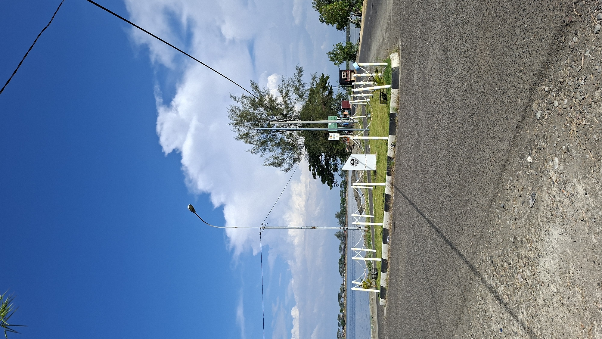 The image depicts a roadside monument on a sunny day with a clear blue sky and scattered clouds, situated near a body of water. The monument, located in the city of Lhokseumawe, Indonesia, is surrounded by a low white fence with black and white striped borders. A single street lamp stands beside the monument, and power lines are visible against the sky. The roadside is paved, with a gravel shoulder in the foreground. In the background, trees and a distant shoreline are visible across the water. There are no people present in the image.