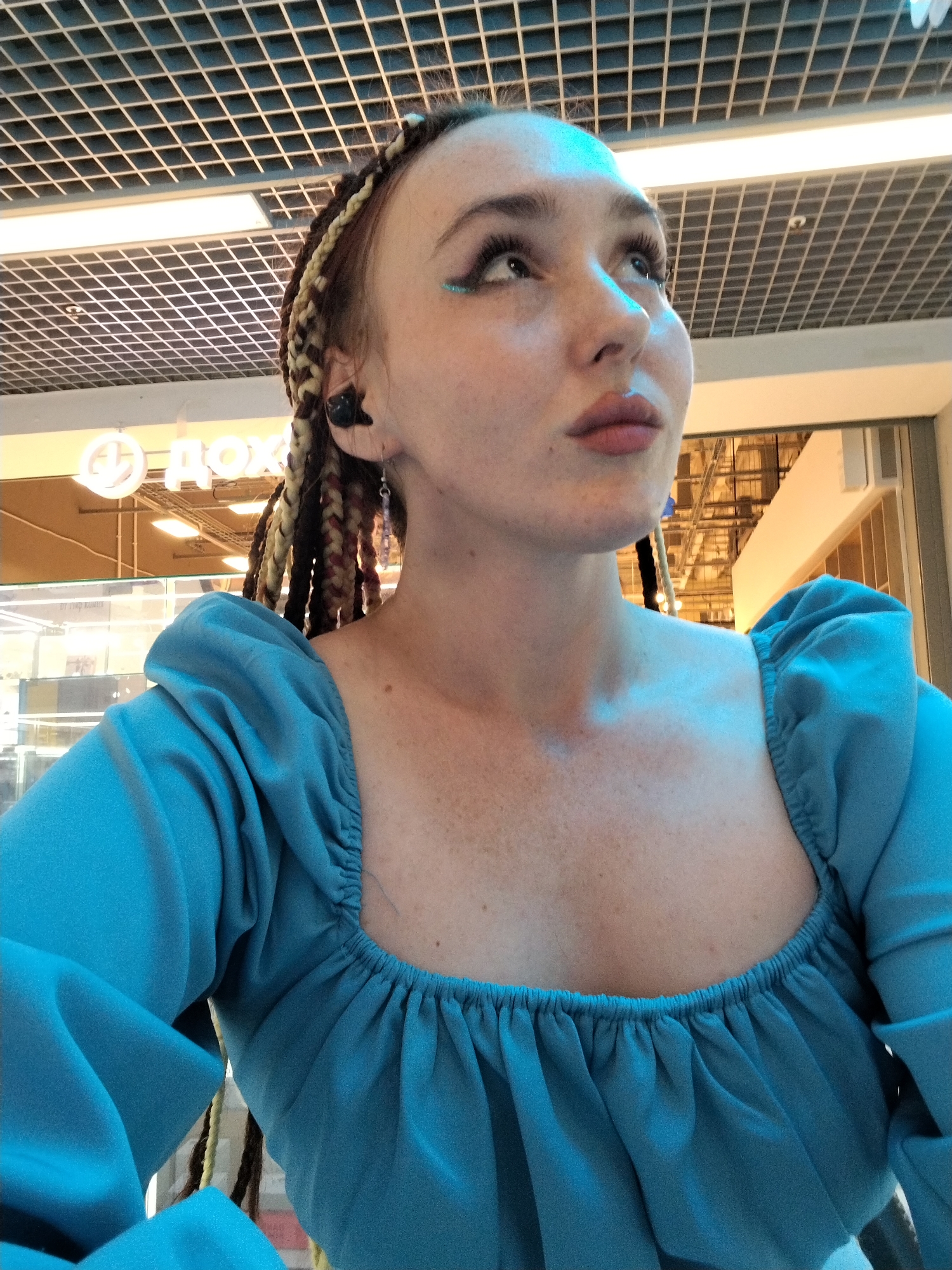 A medium shot depicts a light-skinned person with red hair braided with blonde and brown strands, wearing a blue top with puffy sleeves and a square neckline. They are facing slightly away from the camera and looking up and to the right.
The background indicates an indoor location, possibly a shopping center, identifiable by the overhead grid ceiling with fluorescent lights and the partial view of a storefront sign with Cyrillic lettering “ДОХ”. This sign, in conjunction with other visual cues, suggests the location may be in the city of Izhevsk, Russia. The location is brightly lit. The individual has visible freckles. A black in-ear headphone and earring are visible on the person.