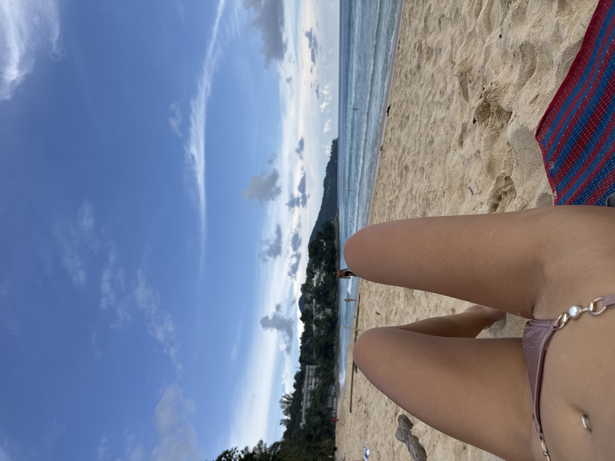 The image shows a first-person perspective of someone lying on a sandy beach in Choeng Thale, Thailand. The foreground is dominated by the person's legs and a portion of their bikini bottoms, with sand visible on their skin. A red and blue striped mat is placed to the right of the legs. In the background, the ocean stretches out under a blue sky with scattered clouds. Waves are breaking on the shore, and a few people are visible in the water and on the beach in the distance. A tree-lined hillside with buildings is visible on the horizon. The scene appears to be a relaxed day at the beach.