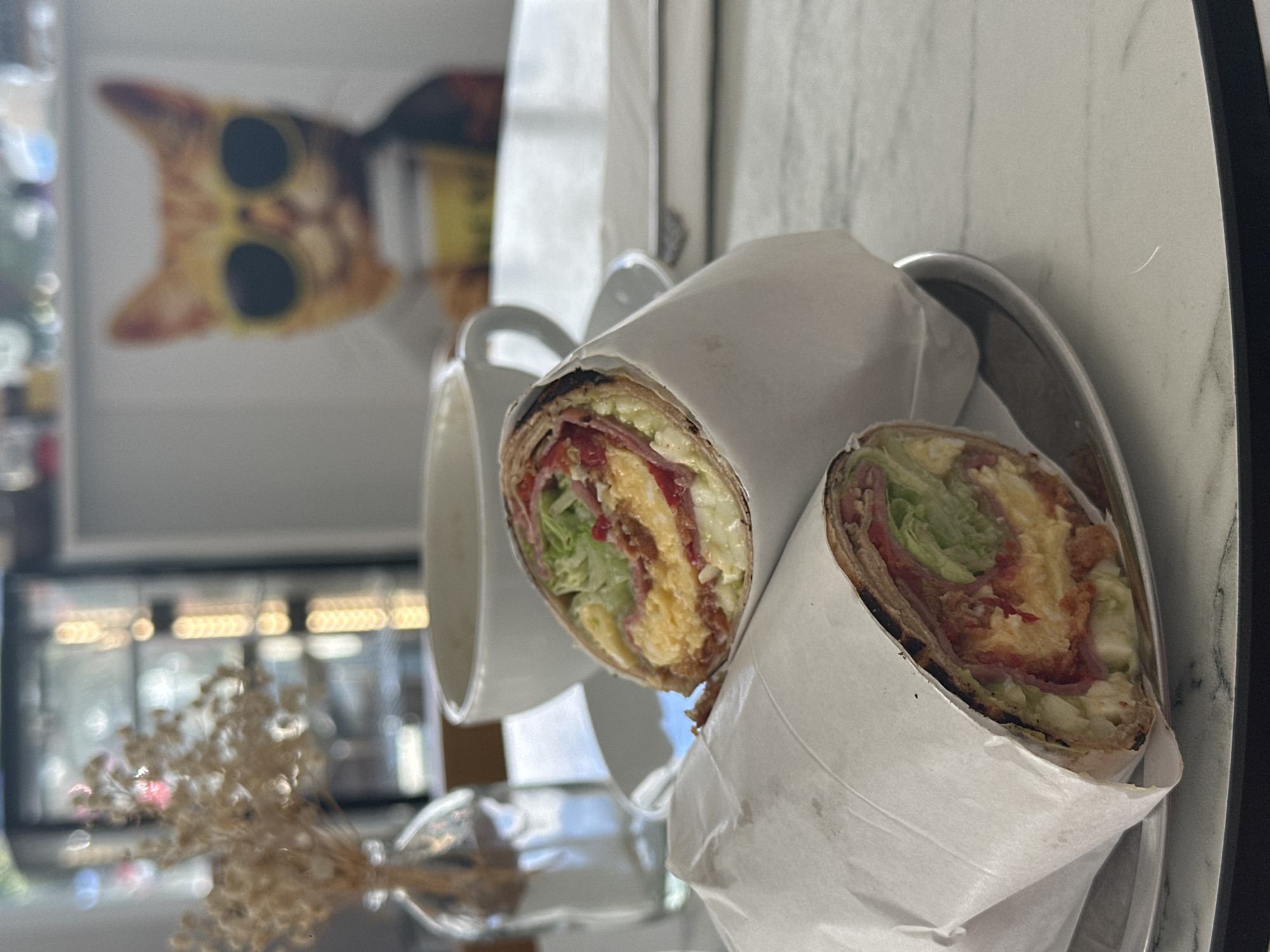A close-up shot shows two wrapped sandwiches on a metal plate. The sandwiches appear to contain lettuce, ham, and a fried component. Behind the plate, a white ceramic mug sits slightly off-center. A blurred image of a cat wearing sunglasses is visible in the background, potentially a piece of artwork or a photograph. In the foreground, a partially visible, vase-like object containing dried plant stems is present. Outside the window is likely the city of Datça, Turkey, based on the prompt. The table is white with a marble-like pattern.