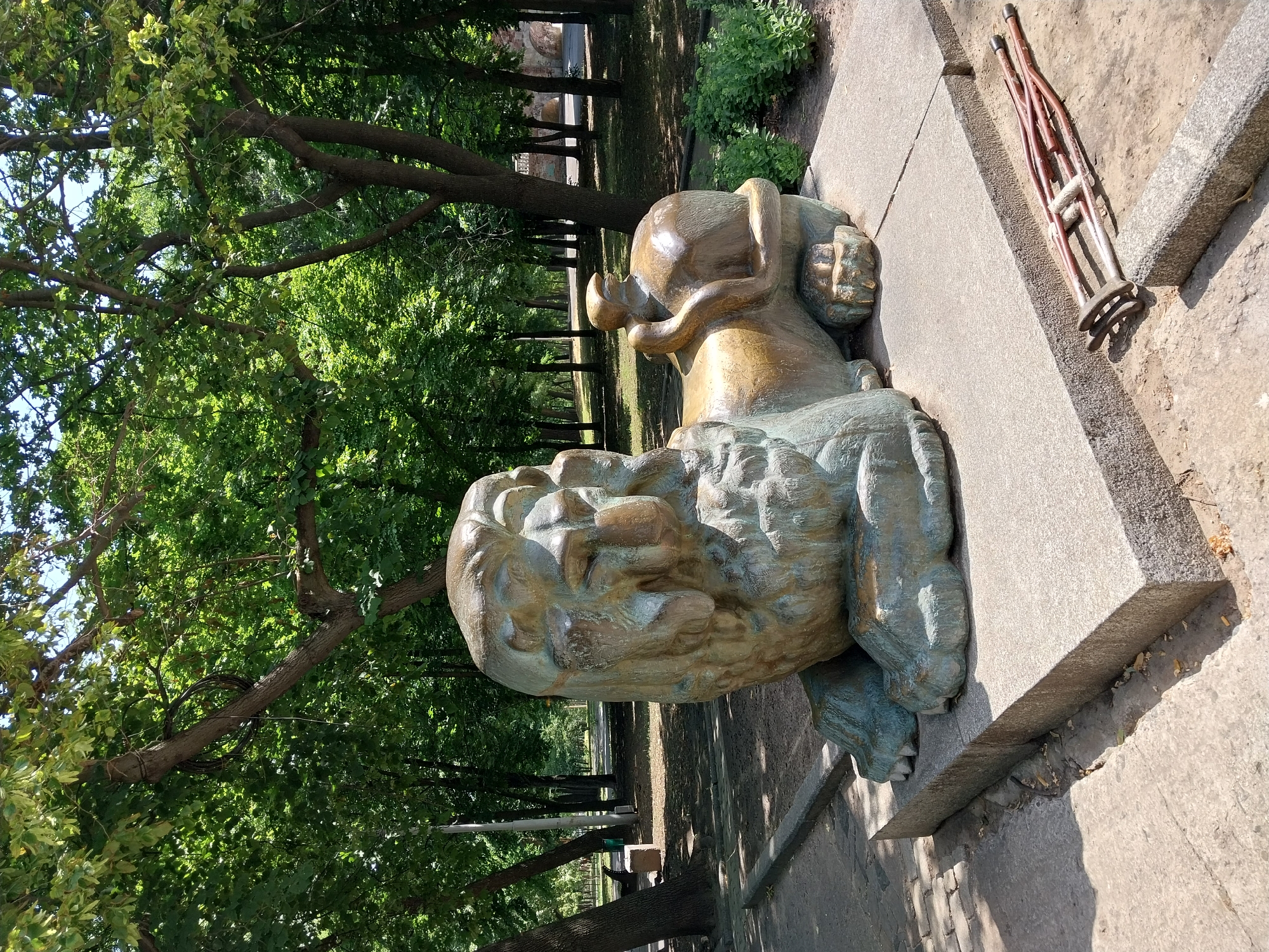 An artistic sculpture of a lion is situated on a concrete pedestal in a park in Dnipro, Ukraine. The lion sculpture, made of bronze or a similar material, exhibits a patina of green and gold. It is depicted in a reclining pose, with its head turned to the left. To the right of the lion, a smaller, rounded object, possibly a representation of an animal or a stylized element, is embraced by the lion's tail.
The sculpture is surrounded by a park environment. Lush green trees with dense foliage create a canopy overhead, dappled sunlight filtering through the leaves. The ground is a mixture of dirt, gravel, and paved pathways. In the foreground, a pair of crutches are placed on the ground beside the pedestal, suggesting a recent presence or a temporary arrangement. In the background, park benches, trees, and pathways are visible, indicating a public recreational space. The overall impression is one of a serene, outdoor setting with an artistic focal point.