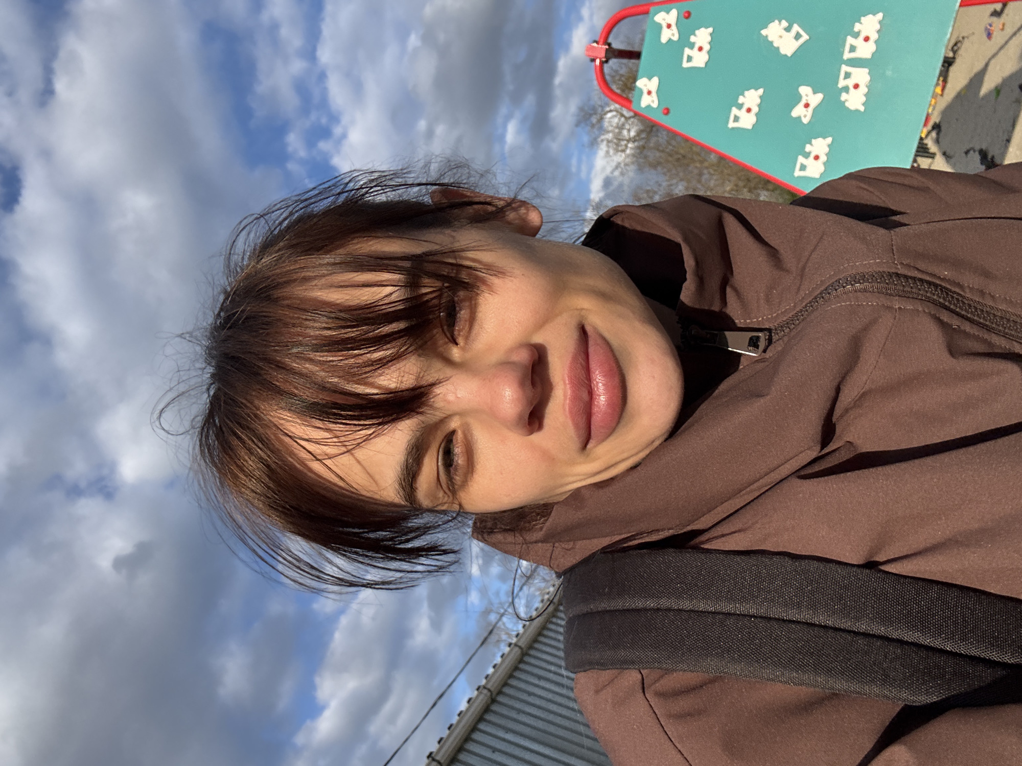 This bright, sunlit image is a close-up selfie of a young person taken outdoors in Rostov-on-Don, Russia. The shot is tilted, presenting the subject's face and upper body angled across the frame from left to right.
The subject has dark brown hair styled with bangs and fair skin, smiling widely with their eyes partially closed, possibly due to the direct sunlight. They are wearing a brown hooded jacket with a visible silver zipper pull on the right side of their chest, and a black backpack strap is visible over their right shoulder.
The background features a vibrant blue sky with scattered white and grey clouds, suggesting a partly cloudy but pleasant day. In the upper right quadrant of the image, a turquoise-colored board with a red metal frame is visible. This board is decorated with several white shapes, possibly resembling gingerbread figures or Christmas trees, each featuring a small red dot, indicative of playground equipment. A corrugated metal fence or roofline can be seen along the bottom left edge of the frame. The overall mood conveyed is one of happiness and enjoyment of the outdoors. No text is visible in the image.