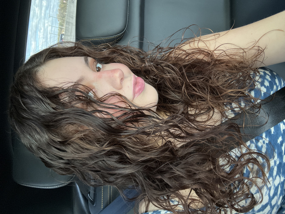 A young woman with long, dark, curly hair and fair skin is positioned in what appears to be a car. She wears a patterned blue and white shirt and a seatbelt is visible across her chest. Her head is tilted to the side, with her gaze slightly upward. The light seems to be coming from the left, casting shadows on her face and hair. The background consists of the car's interior.