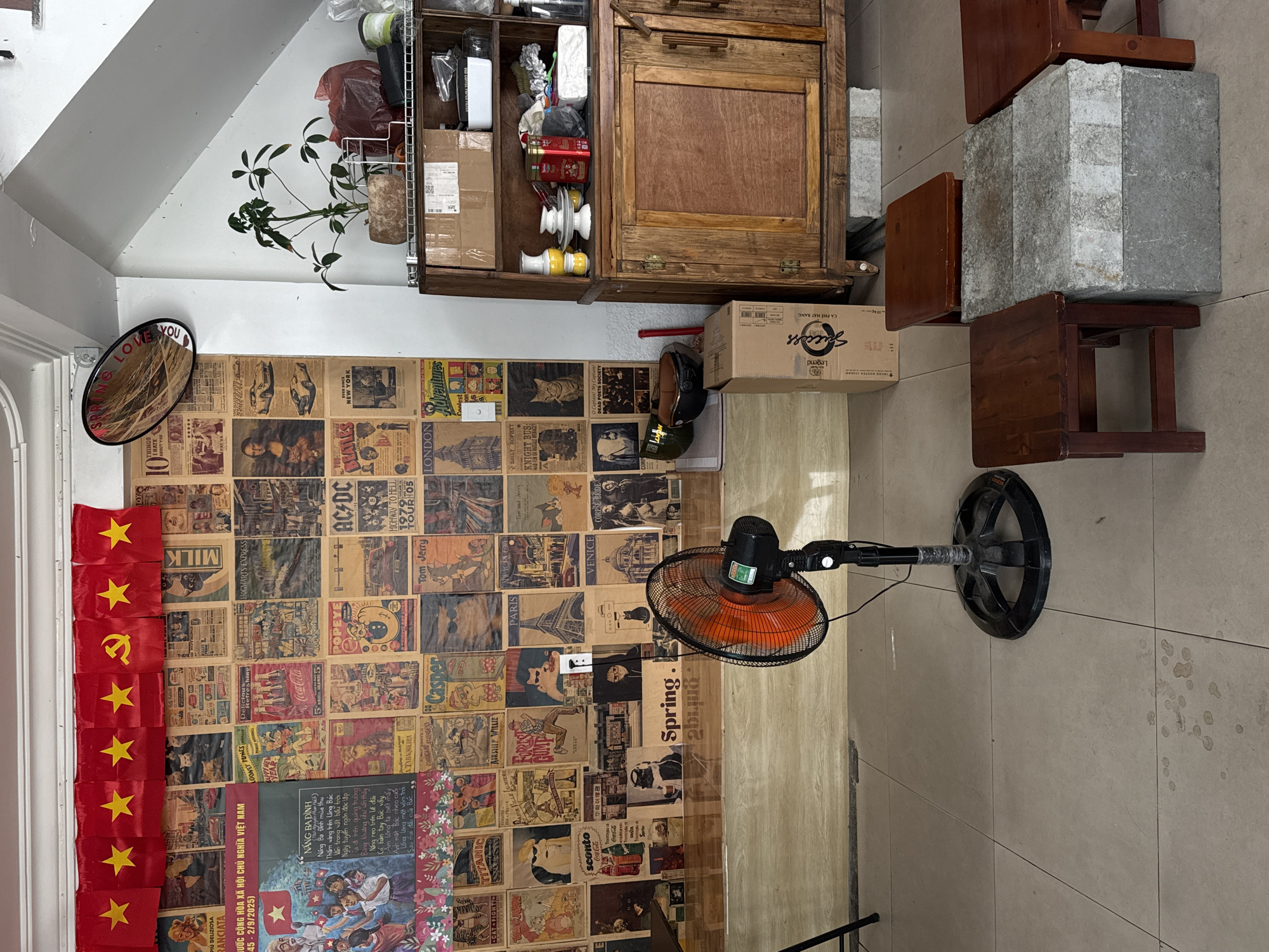 The image shows an interior space in what appears to be a shop or small business in Đà Nẵng, Vietnam. A wall is almost entirely covered with vintage newspaper clippings and posters. To the left, a Vietnamese flag hangs vertically. A mirror with the words "SPRING LOVE YOU" is mounted on the wall. A wooden cabinet is filled with various items, including boxes, bottles, and containers. A floor fan with an orange blade stands on the tiled floor. The presence of newspapers, the cabinet, and the fan suggest a lived-in and functional space.