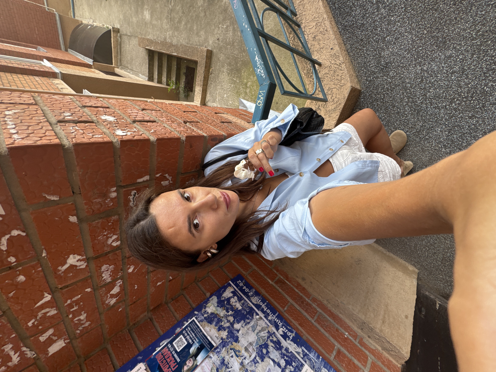 A woman is taking a selfie outdoors, holding an ice cream cone in her left hand. She is wearing a light blue button-up shirt, white eyelet shorts, and tan slip-on shoes. She has large hoop earrings and several rings on her fingers. A black purse is slung over her shoulder.
The background shows a brick wall with peeling posters, a concrete walkway with steps, and a blue metal railing. The scene appears to be in a courtyard or alleyway in the city of Chaikovsky, Russia. The lighting suggests it is daytime.