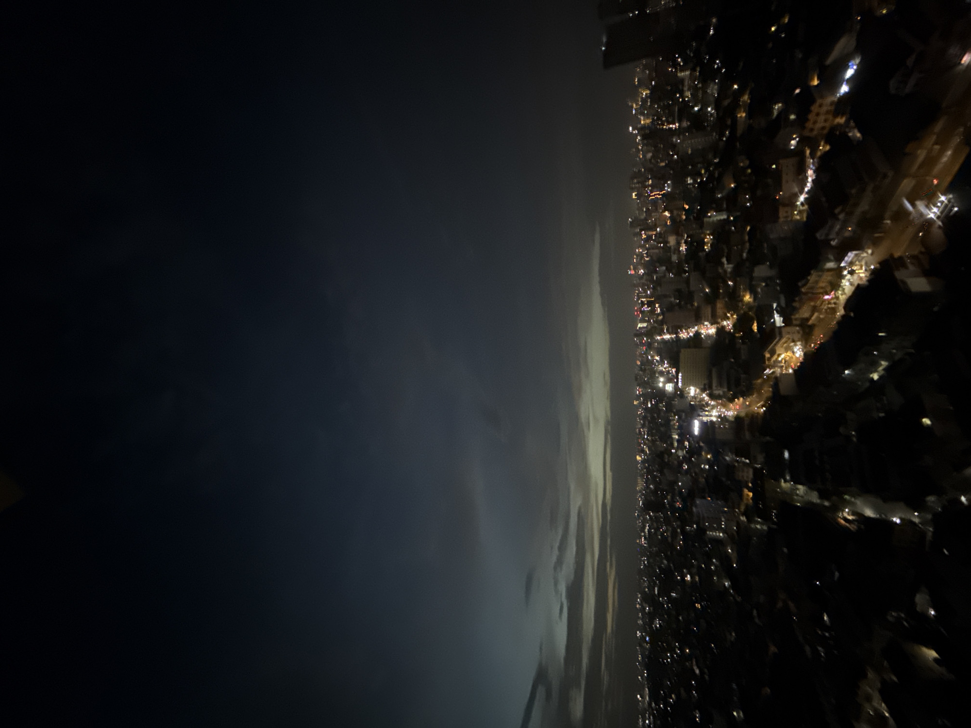 The image captures a nighttime aerial view of a dense urban landscape, likely at dusk or pre-dawn, given the dark, cloudy sky with faint illumination on the horizon. Numerous city lights twinkle below, indicating a sprawling metropolis. Roads are visible, lined with streetlights and illuminated by building lights. The architecture appears to be a mix of low-rise buildings and some taller structures, particularly on the right side of the frame. The overall impression is that of a vibrant, active city at night. This scene is consistent with the urban environment of Khan Mean Chey, Cambodia, known for its dense population and commercial activity. There are no discernible people or specific actions captured in this wide shot.