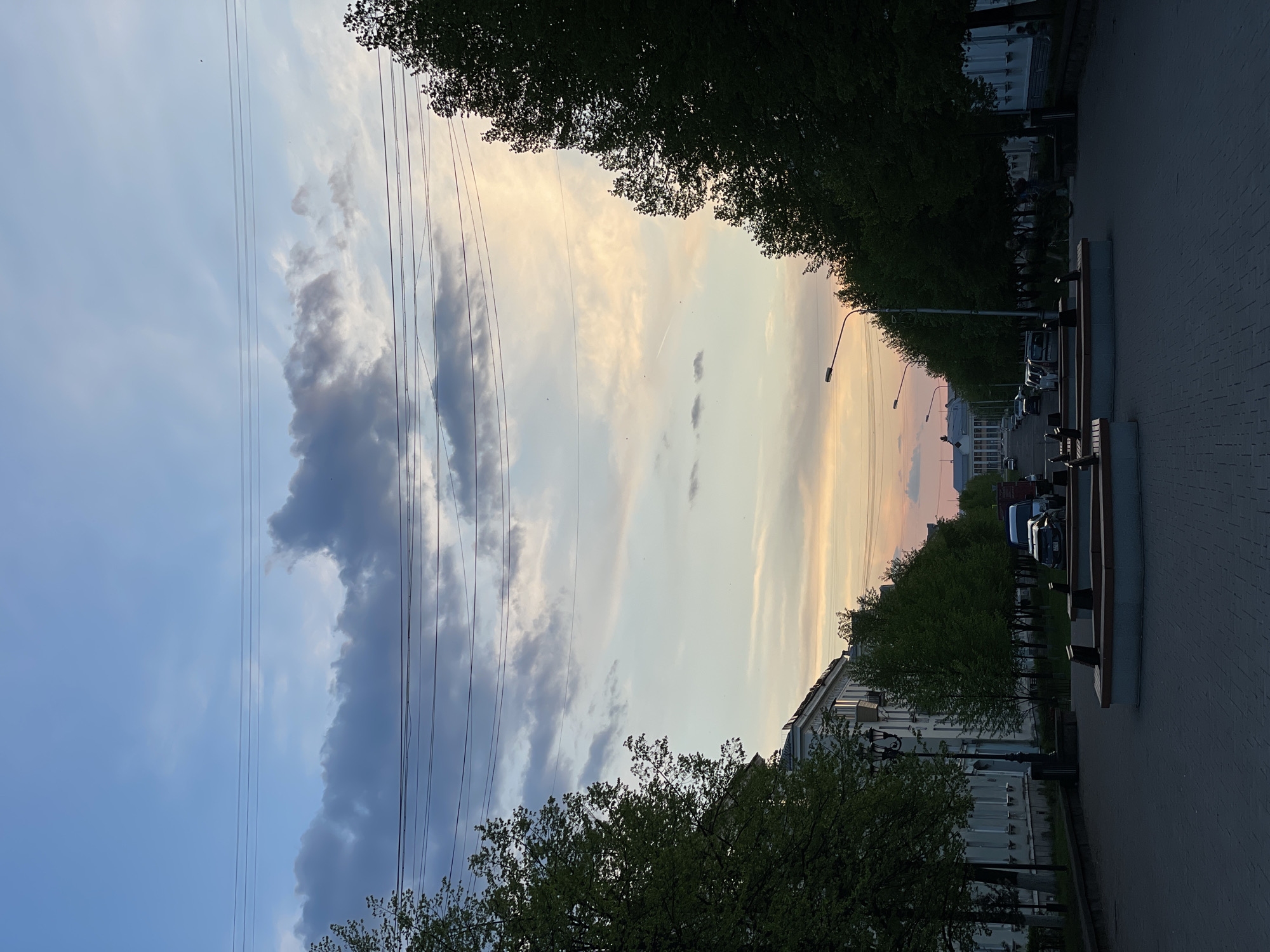 A horizontal outdoor shot shows a street in Zheleznogorsk, Russia, with trees lining both sides of the sidewalk.
The sky is multi-colored, transitioning from blue with dark clouds on the left to a light yellow/orange hue towards the right. Power lines are visible crossing the sky.
The street has a brick sidewalk with benches and streetlights. Cars are parked along the right side of the street, near the building.