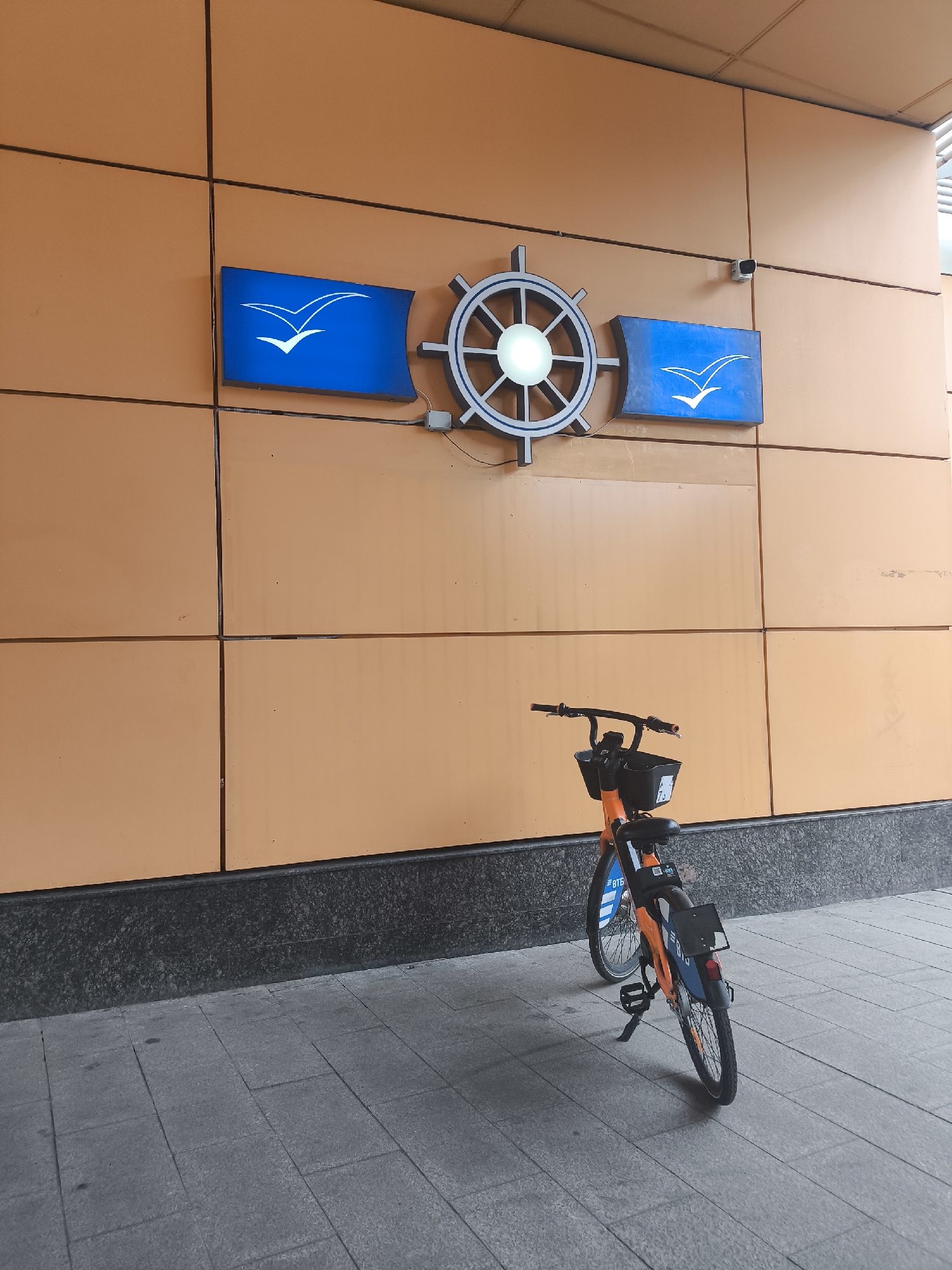The image shows a bicycle parked on a gray-tiled sidewalk in front of a wall consisting of brown panels. The bicycle is orange and blue, with a black basket on the handlebars. Above the bicycle on the wall is a logo that features two blue rectangular signs, each with a white stylized image of a bird in flight. A white ship's wheel is located between the signs. A security camera is positioned on the wall to the right of the ship's wheel.