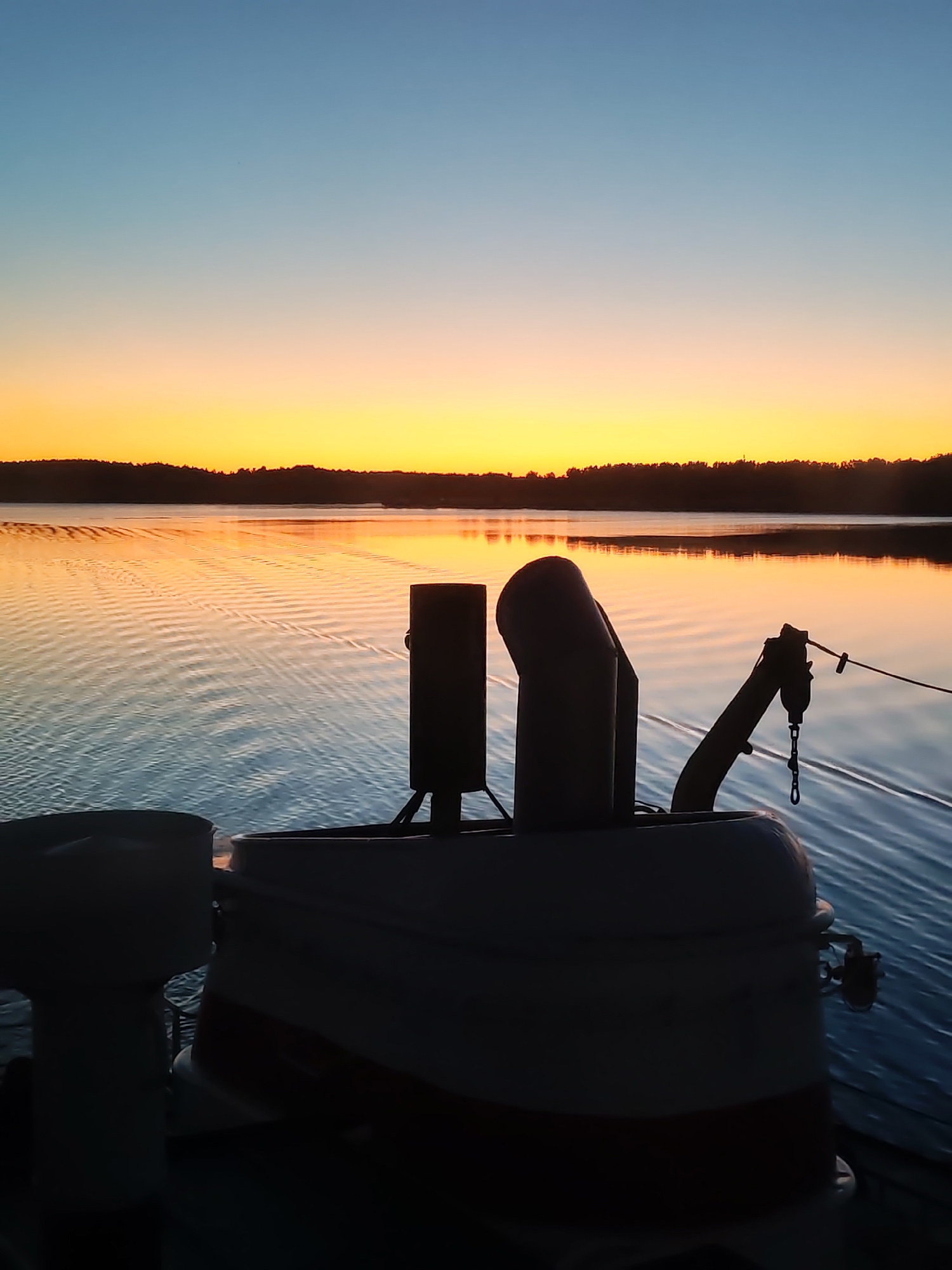 An image captured from the deck of a boat depicts a sunrise or sunset over a body of water with a distant landmass. The sky exhibits a gradient, transitioning from a light blue at the zenith to vibrant orange and yellow hues at the horizon. A dark, silhouetted line of trees or dense vegetation forms the distant shoreline, separating the sky from the water. The water's surface reflects the sky's colors, displaying orange and yellow tones near the horizon that fade into darker blues and grays closer to the foreground. Distinct ripples and wave patterns are visible on the water's surface, particularly in the middle ground. In the immediate foreground, various dark, silhouetted structures belonging to the boat are visible. These include a partial cylindrical structure on the far left, two prominent pipe-like or vent-like structures in the center (one vertical, one angled), and an articulated arm or crane-like mechanism with a hook and chain hanging from it on the right. The upper part of the boat's hull or deck railing is also visible in silhouette below these structures, with a faint lighter band discernable on the central boat structure. No individuals are present in the scene. The location is Unknown, Unknown.