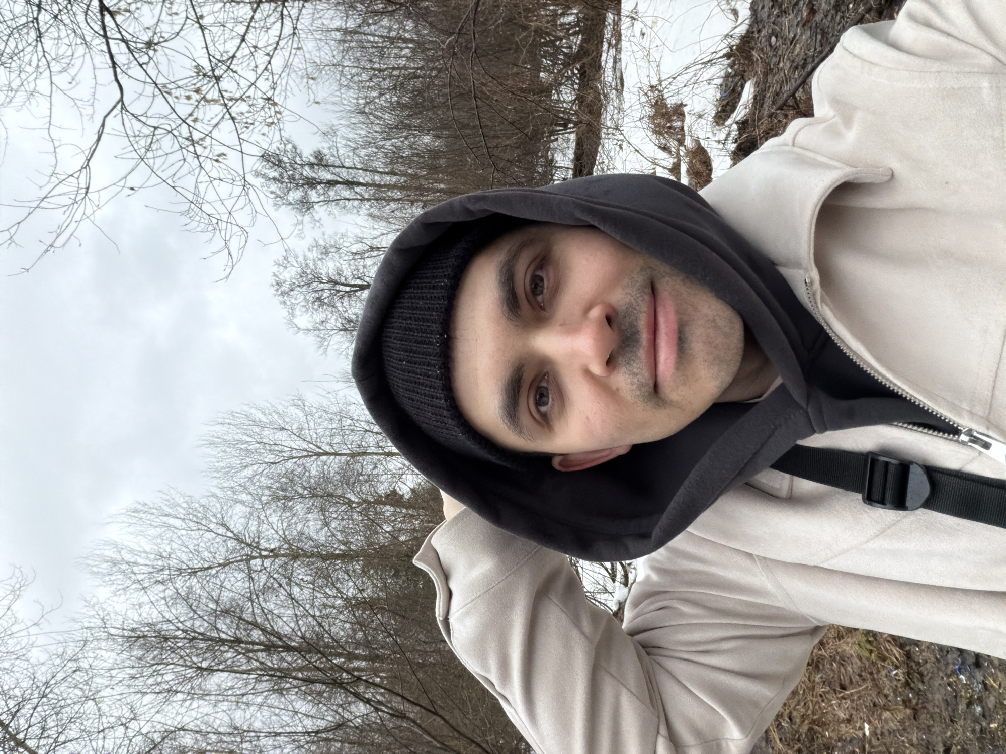 A selfie depicts a man in an outdoor setting. He is wearing a black beanie and a black hoodie under a beige jacket. The jacket is partially zipped, revealing a black strap across the chest. The background consists of trees with bare branches under an overcast sky. The ground appears to be covered with mud and some snow. The location is possibly near a wooded area in a city with bare trees. The exact location in Izhevsk, Russia cannot be determined from the image.