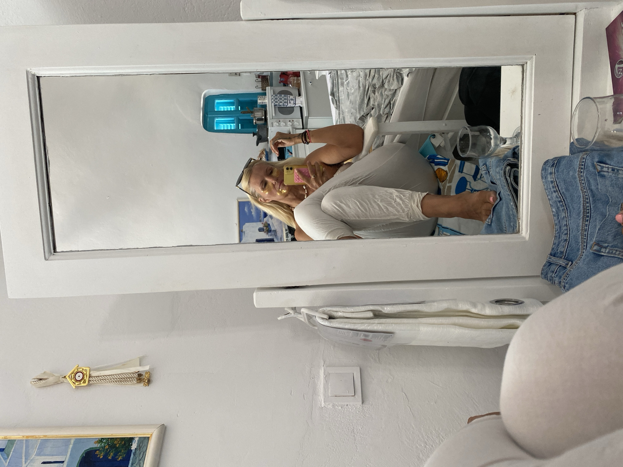 A woman is taking a selfie in a mirror in a room. She is sitting in a chair, wearing white pants and a white top. She has gold under-eye patches on her face and is holding a yellow smartphone. In the background, there is a bed, a microwave, and a blue window. On the wall, there is a painting and a decorative hanging. There are also clothes and a glass on the floor in front of the mirror. The scene appears to be in a room in Ia, Municipal Unit, Greece, given the characteristic white walls and blue accents.