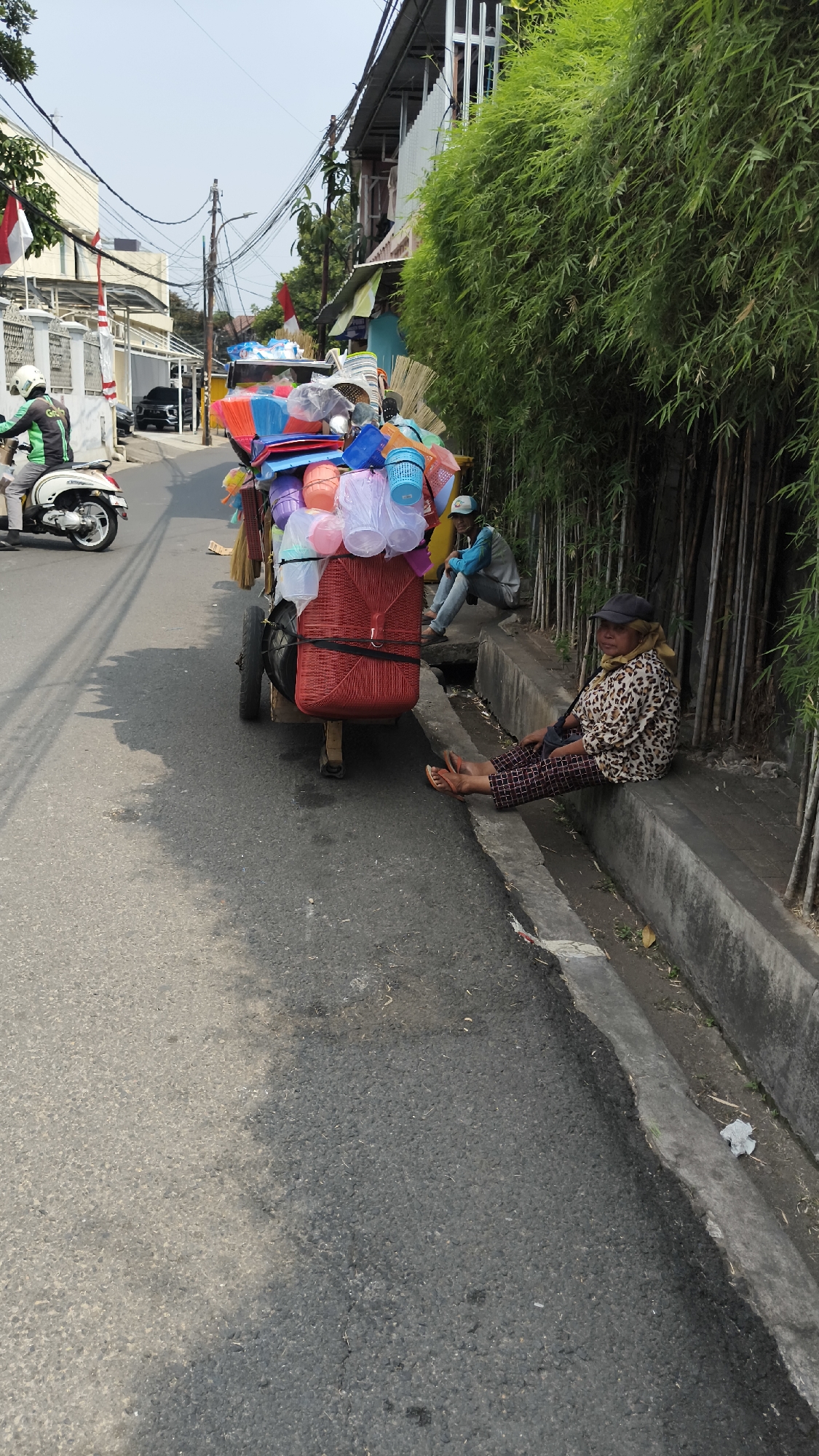 The image depicts a street scene in South Jakarta, Indonesia. A cart laden with colorful plastic household items, including buckets and bowls, is parked on the side of the asphalt road. A woman wearing a leopard-print shirt, dark checkered pants, a black cap, and sandals is sitting on a concrete curb next to the cart, facing the camera. Another person, wearing a blue long-sleeved shirt and jeans, is seated further down the curb, partially obscured by the cart. A motorcycle with a rider wearing a green uniform is visible on the left side of the road, moving away from the camera. The environment includes a dense bamboo growth on the right side of the curb and buildings with visible power lines in the background. The sky is clear and bright, indicating daytime.