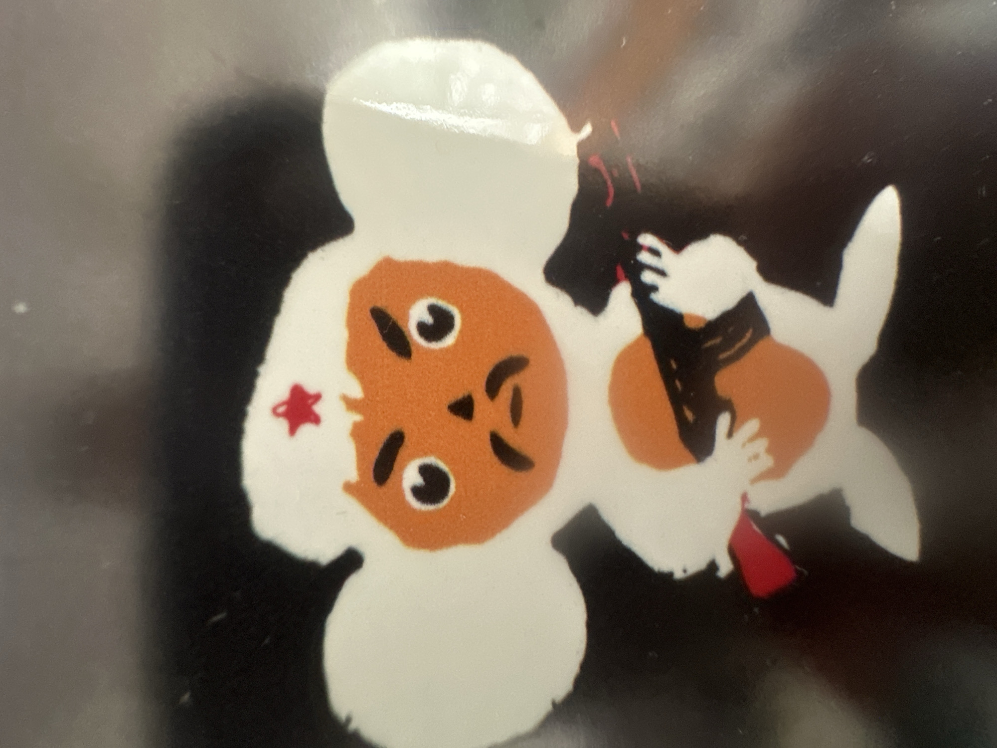 This is a close-up, slightly angled photograph of a small, flat, decorative object with a cartoonish design. The design features two stylized figures appearing to interact. One figure, in the foreground and more prominent, has a round, orange face with simple black eyes, eyebrows, and a mouth. It has a large white head covering and a white body. A small red star is visible on its left cheek. The other figure, to the right, is depicted in white and is interacting with the first figure, appearing to hold its orange head with its white hands. This second figure has an orange circular object near its chest and is mostly obscured by the first figure. The background is dark and appears to be reflective, possibly a surface like glass or polished metal, with some soft, diffused lighting creating a shadow on the left side. The overall aesthetic is playful and childlike. There is no visible text in the image. The context of Belgrade, Serbia, does not provide specific visual cues within this abstract and stylized image.