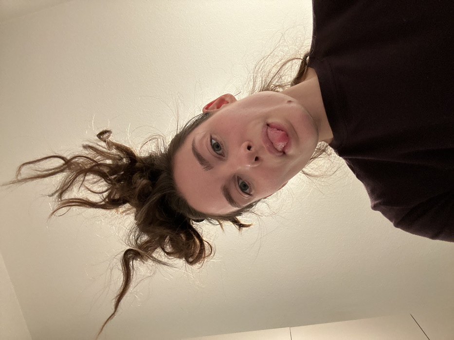 An upward-facing, low-angle shot captures a single young adult female from the neck up, appearing inverted relative to the camera's orientation. The individual has fair skin, light-colored eyes, and brown hair pulled into a messy bun at the back of her head, with several strands cascading downwards due to gravity. She wears a dark, long-sleeved top, possibly dark purple or maroon. Her eyes are open and directed towards the camera. Her tongue is extended from her mouth, with its tip forming a slight V-shape. The background is a plain, light-colored (off-white) wall or ceiling, with a subtle vertical line visible on the far right edge of the frame. Location: City of Unknown, Unknown.
