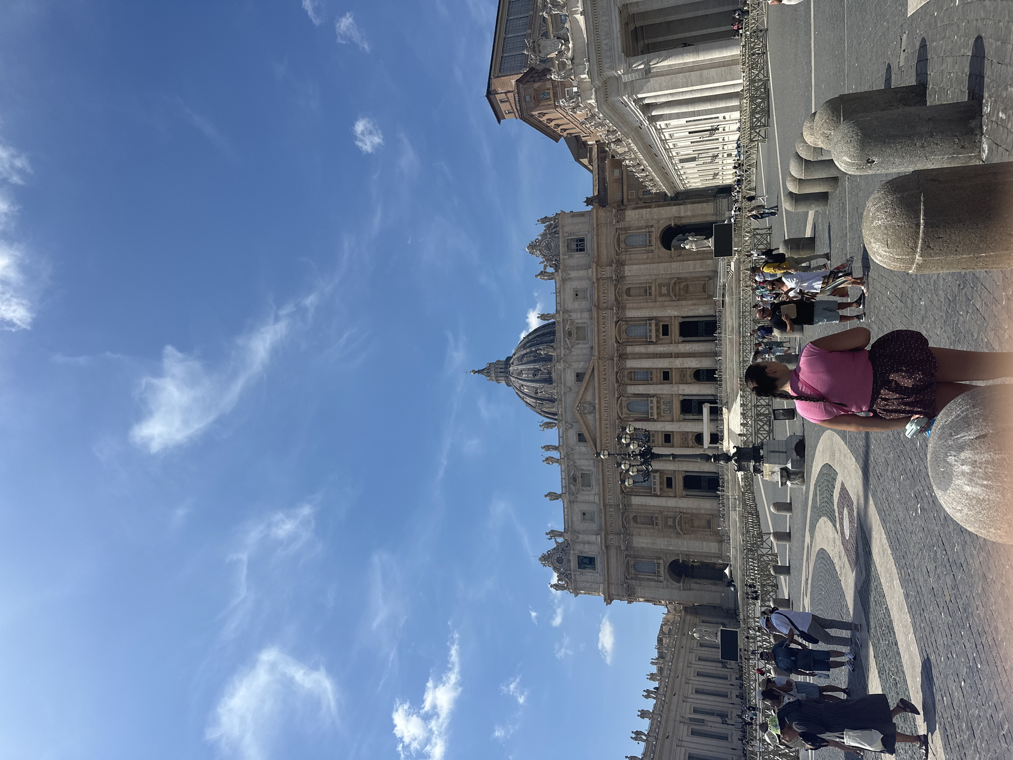 This is a vertical, wide-angle, eye-level, outdoor, daytime photograph taken in St. Peter's Square, Città del Vaticano, Vatican City. The sky is blue with scattered white, wispy clouds. St. Peter's Basilica is the dominant structure in the background, featuring its iconic dome and classical facade. To the right of the Basilica, a colonnade with marble columns is visible, part of the Vatican's architecture. The foreground is paved with cobblestones arranged in a pattern. Several people are present in the square, appearing as tourists. Some are walking, others are standing, and some are engaged in conversation. A woman in a pink shirt and patterned skirt is in the lower right of the frame, facing away from the camera. A group of people are standing near the center, appearing to be taking photos. In the mid-ground, a series of stone bollards are lined up. The overall scene depicts a bustling tourist area with significant architectural landmarks.