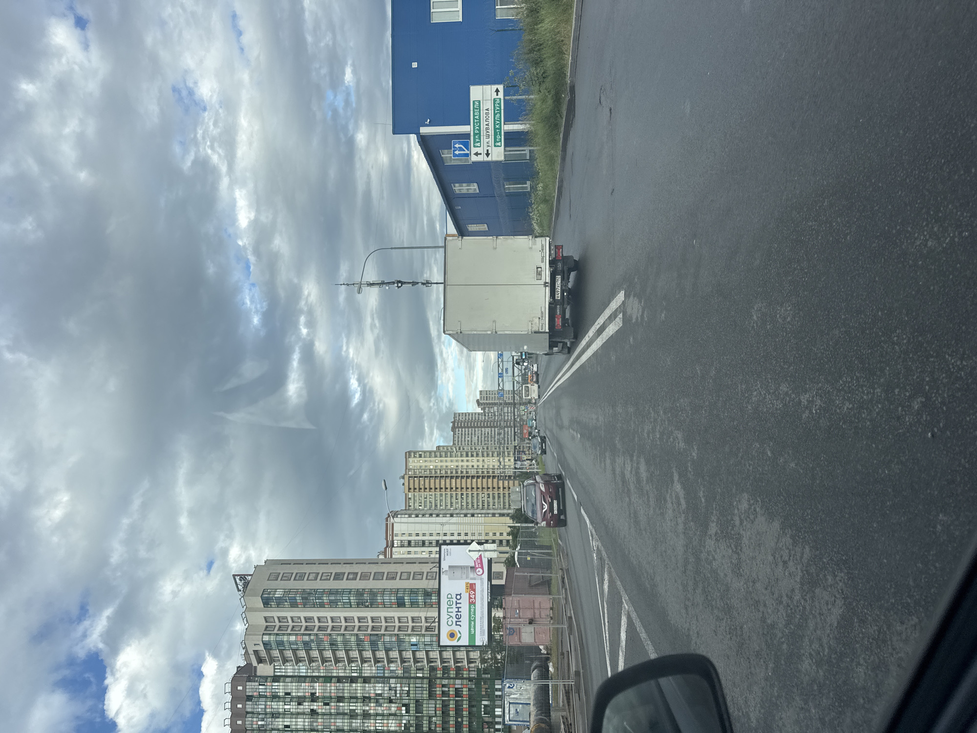 This is a vertical, eye-level, outdoor shot of a street scene in an urban environment. The perspective is from a vehicle, looking forward and slightly to the left. A dark asphalt road with white lane markings occupies the right half of the frame, showing some wet patches and debris. On the left side of the road, there are several tall, modern buildings, most of which are light-colored with many windows. The sky is filled with dramatic, heavy gray clouds interspersed with patches of blue sky, suggesting an overcast day with potential for rain.
In the foreground of the left side, a white box truck is visible, with the license plate "K497xC1Z" and the brand "IVECO" partially visible. Behind the truck, a blue building with white window frames is situated, with a street signpost next to it. The street signs are in Russian, indicating directions to "ул. РУСТАВЕЛИ" (Rustaveli St.), "ул. ШУВАЛОВА" (Shuvalov St.), and "пр-т КУЛЬТУРЫ" (Culture Ave.). Further down the street, more apartment buildings of varying heights and colors line the road, and there is some traffic including cars and a bus. A large advertisement billboard for "супер лента" (super tape/ribbon) with a price indication of "349" and a "-45%" discount is also visible on the left. The overall impression is a somewhat gray and utilitarian urban landscape.